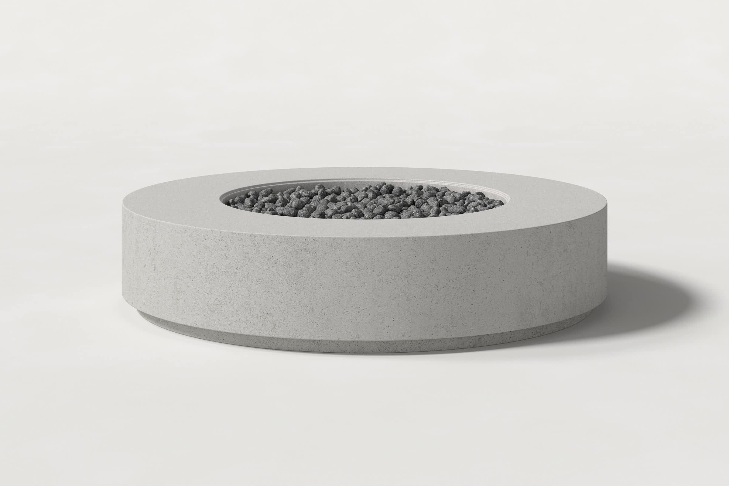 Low cylindrical soft grey concrete fire pit with thick walls and a central circular well filled with dark volcanic rocks. Side view on a white background with a shadow to the right.