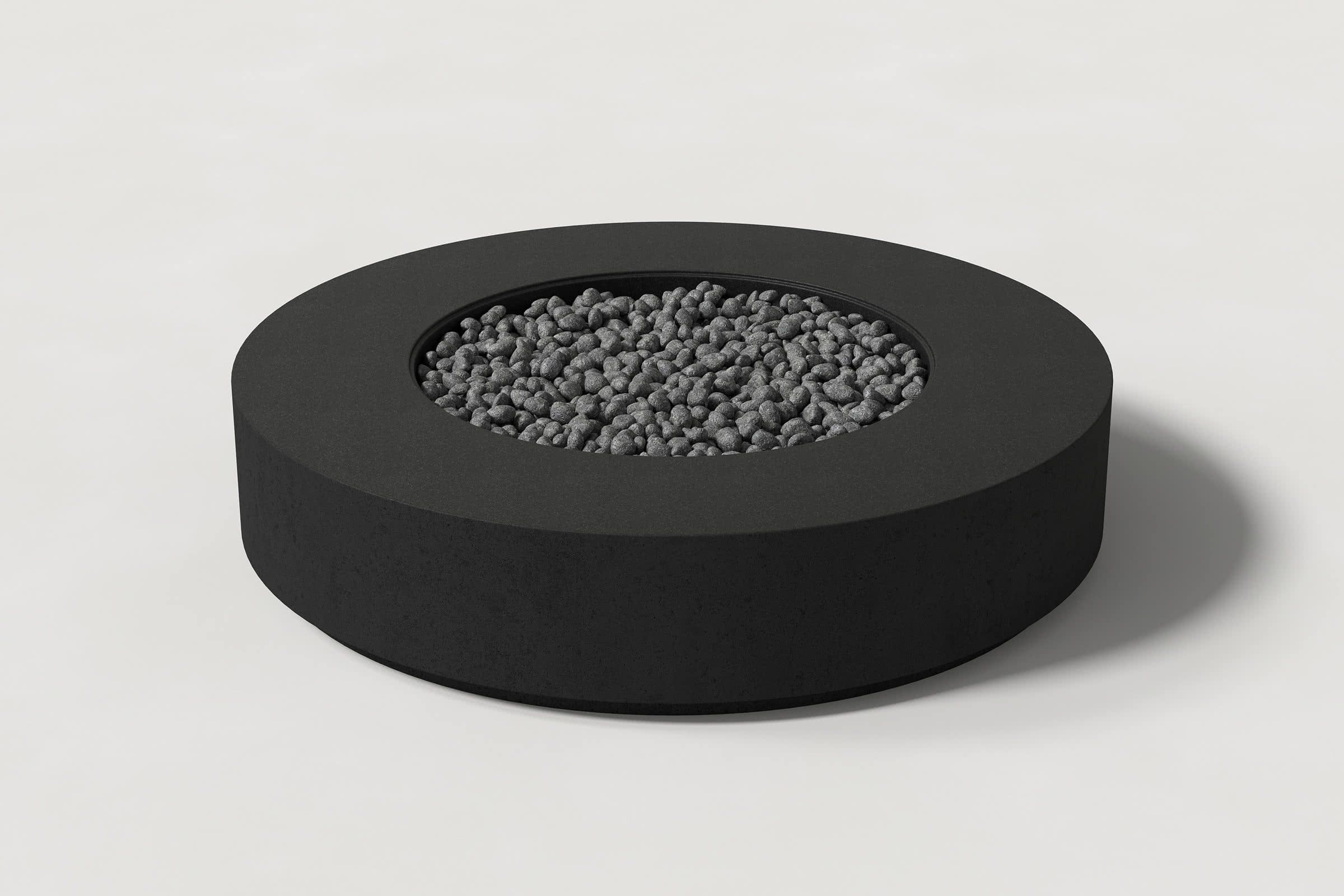 Low cylindrical jet black concrete fire pit with thick walls and a central circular well. Mid-range angle shows the well filled with volcanic rocks against a white background.