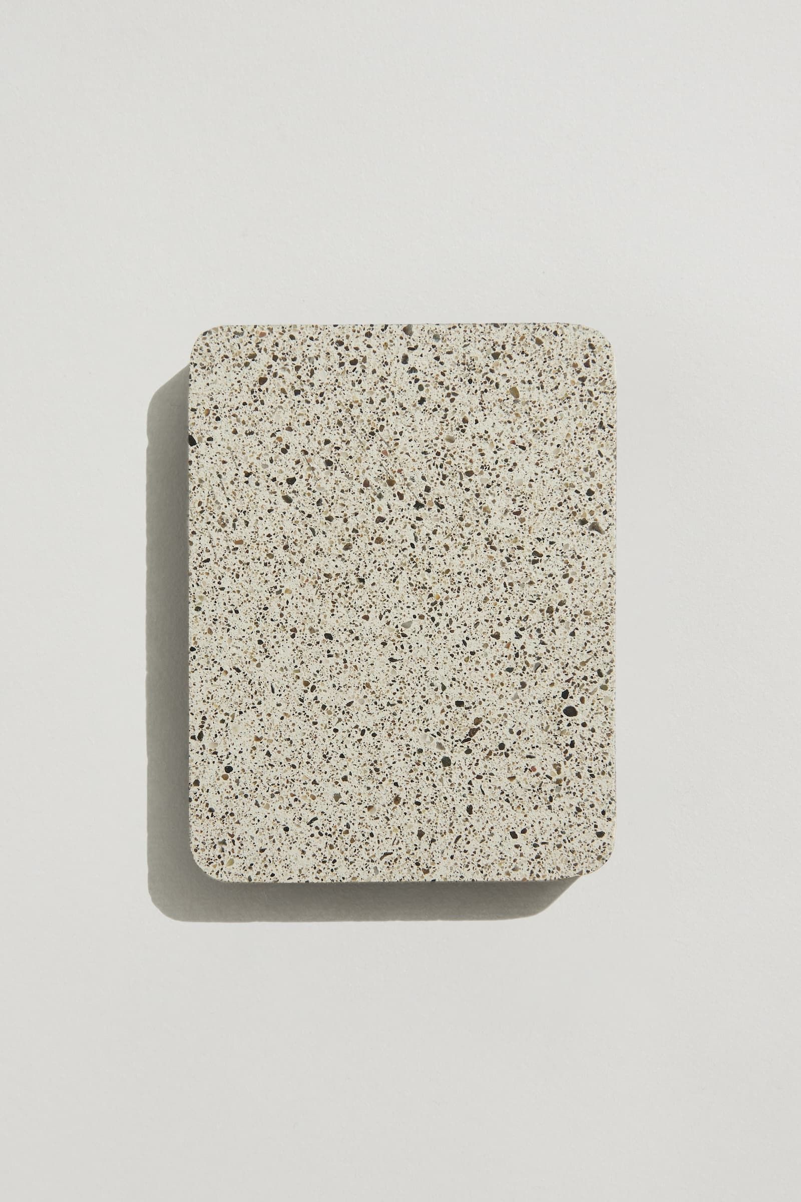 A rectangular sample of white concrete with small black and brown aggregate speckles, and rounded edges. Photographed on a white background with a cast shadow