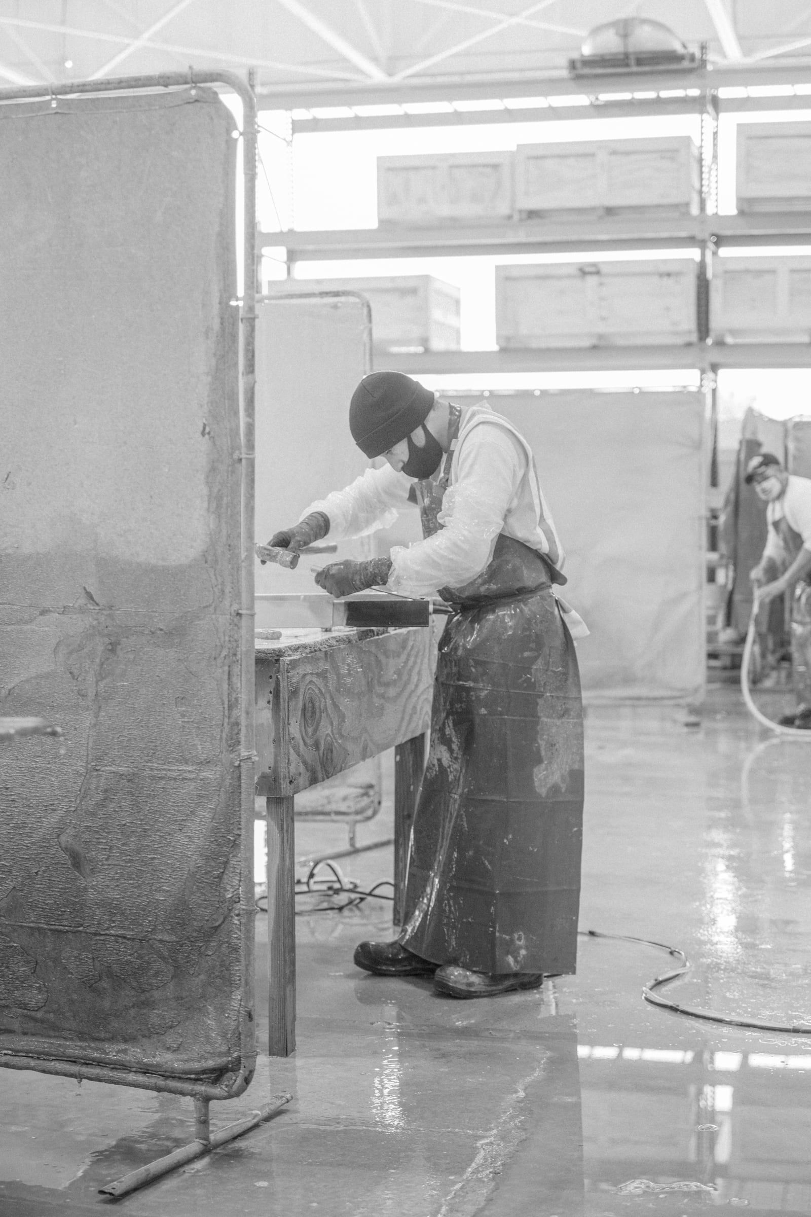 Concrete crafter in protective gear using a hammer to shape a concrete slab in a dusty worksite environment, shown in greyscale.