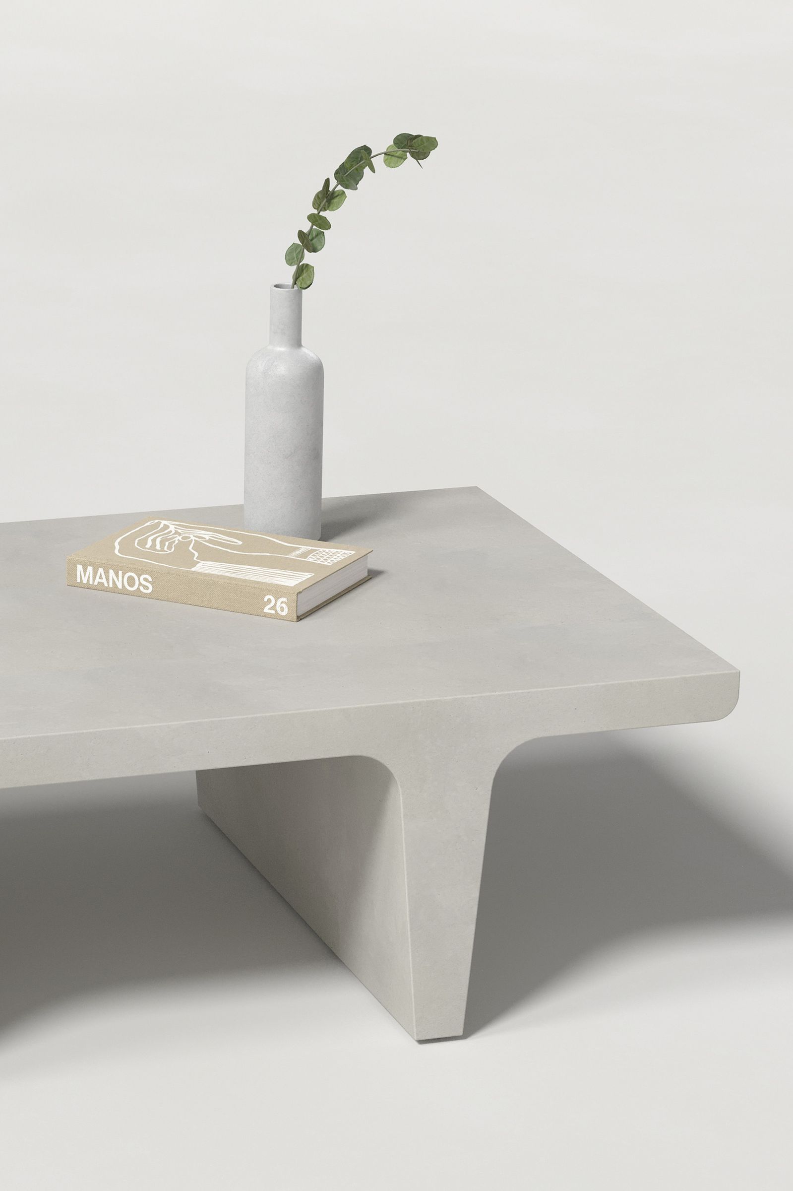 Close-up of a rectangular light grey concrete coffee table with sculptural double tee legs, styled with a neutral-toned book and a slender vase holding a eucalyptus, set against a clean white background.