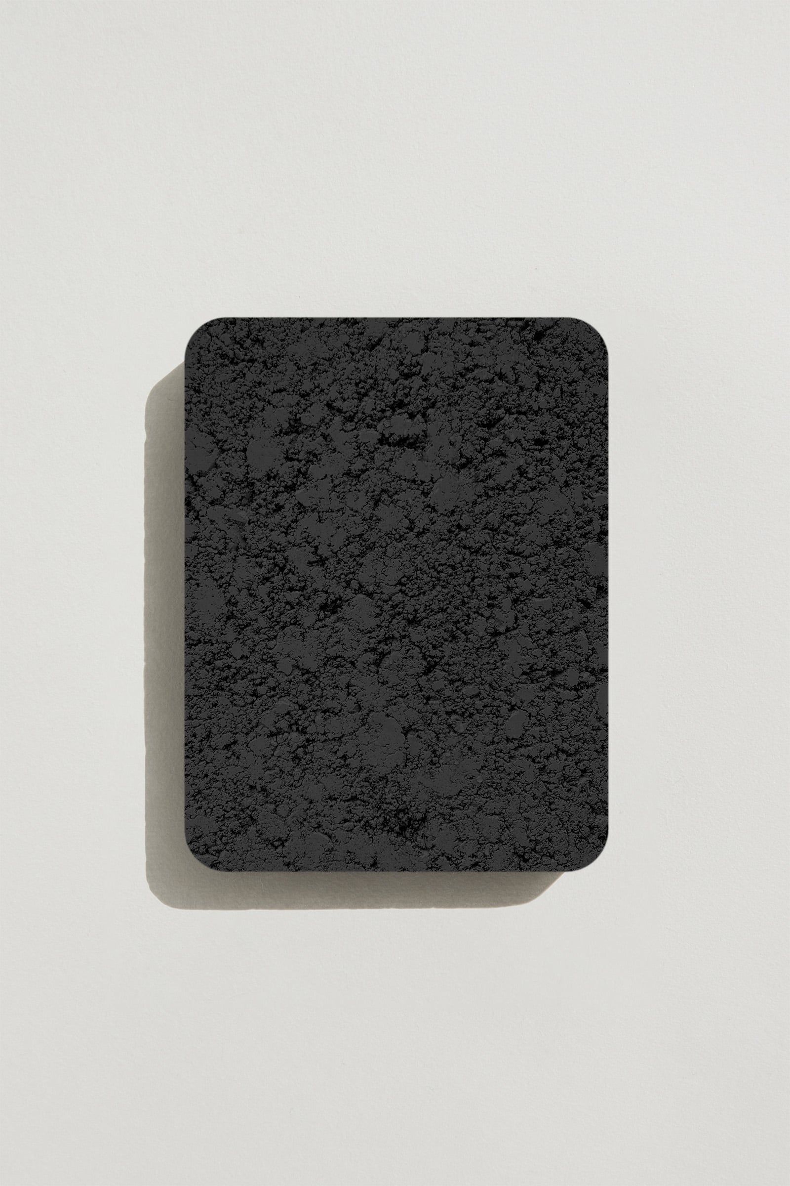 A rectangular sample of rough charcoal concrete with a natural, pitted surface texture. Photographed on a white background with a cast shadow.