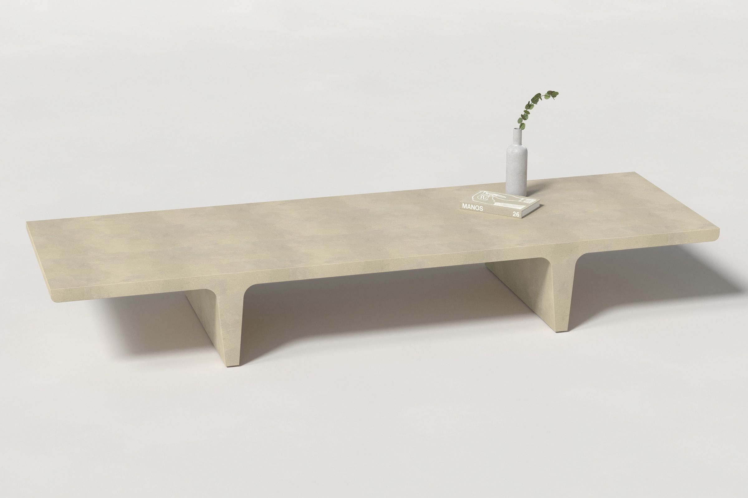 A long rectangular light beige concrete coffee table with sculptural double tee legs, styled with a neutral-toned book and a slender vase holding a eucalyptus, set against a clean white background.