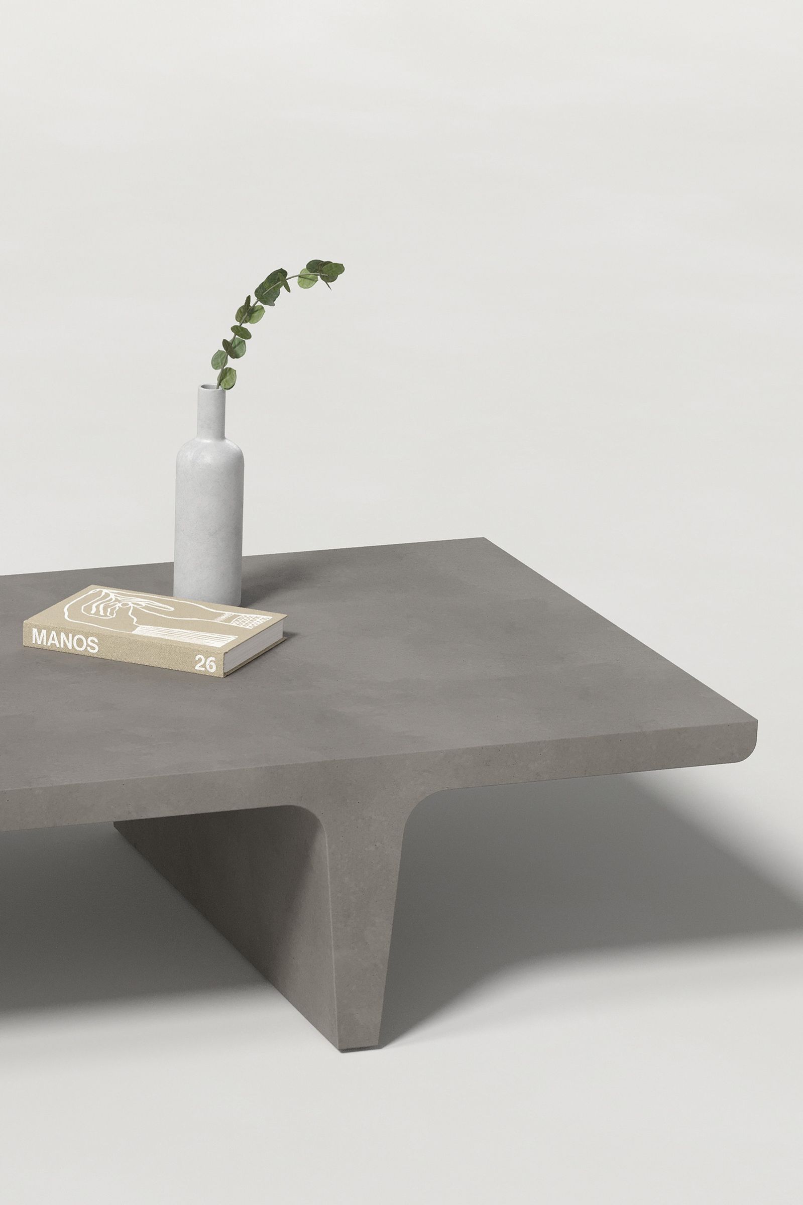 Close-up of a rectangular dark grey concrete coffee table with sculptural double tee legs, styled with a neutral-toned book and a slender vase holding a eucalyptus, set against a clean white background.