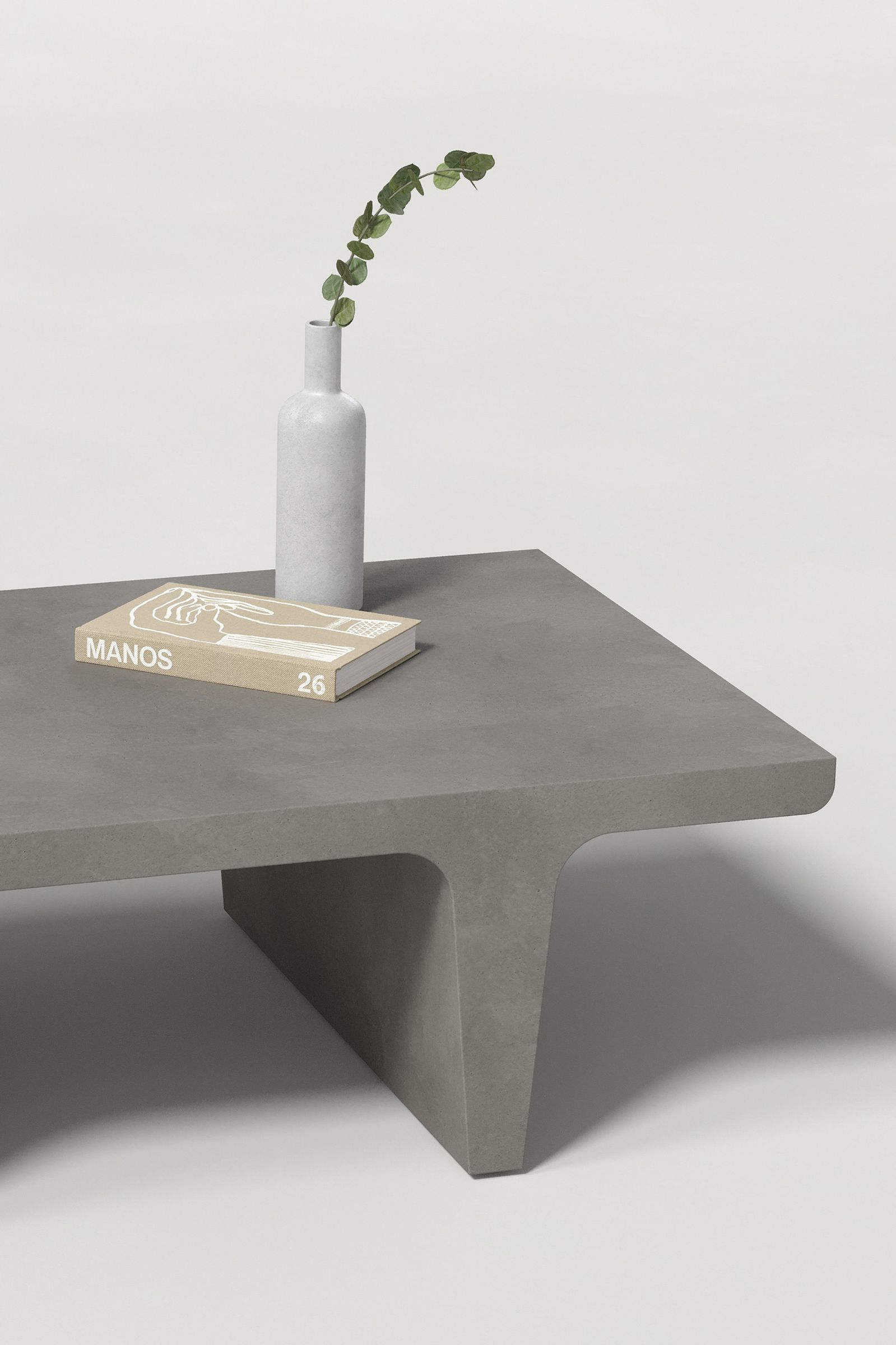 Close-up of a rectangular dark grey concrete coffee table with sculptural double tee legs, styled with a neutral-toned book and a slender vase holding a eucalyptus, set against a clean white background.