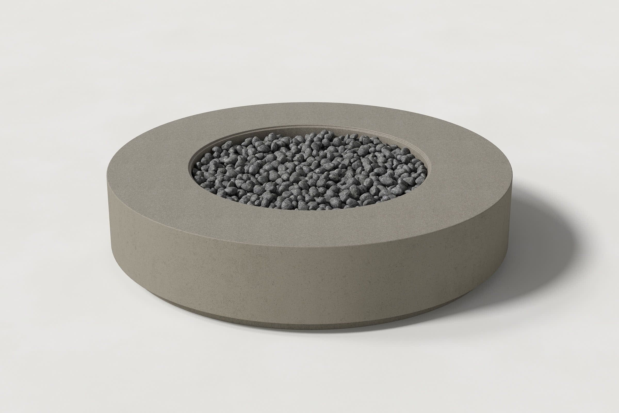 Low cylindrical dark grey concrete fire pit with thick walls and a central circular well. Mid-range angle shows the well filled with volcanic rocks against a white background.