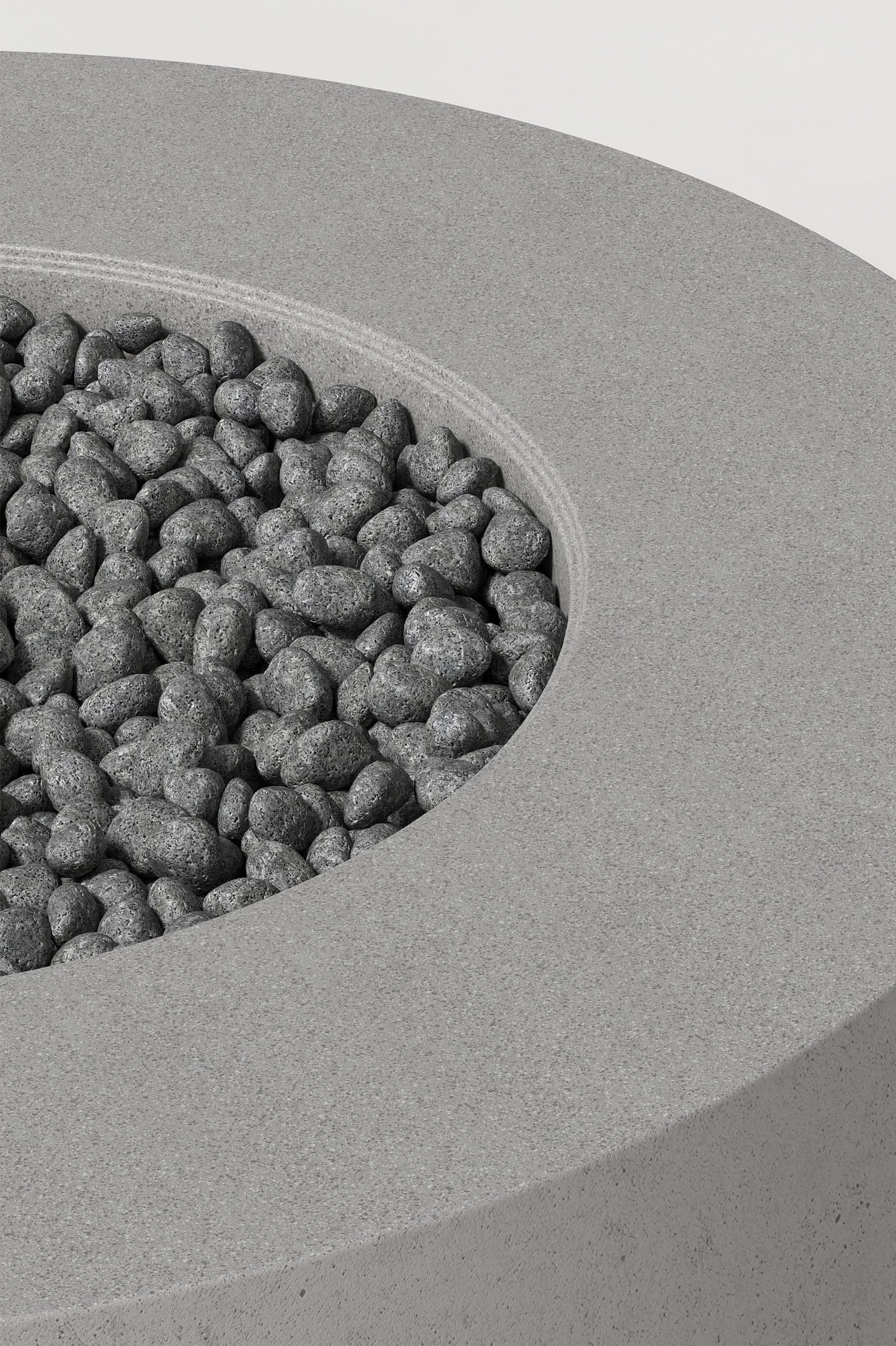 Close-up photograph of a cylindrical grey concrete fire pit with thick walls and a well filled with volcanic rocks. Showcases a smooth texture and precise edge design.