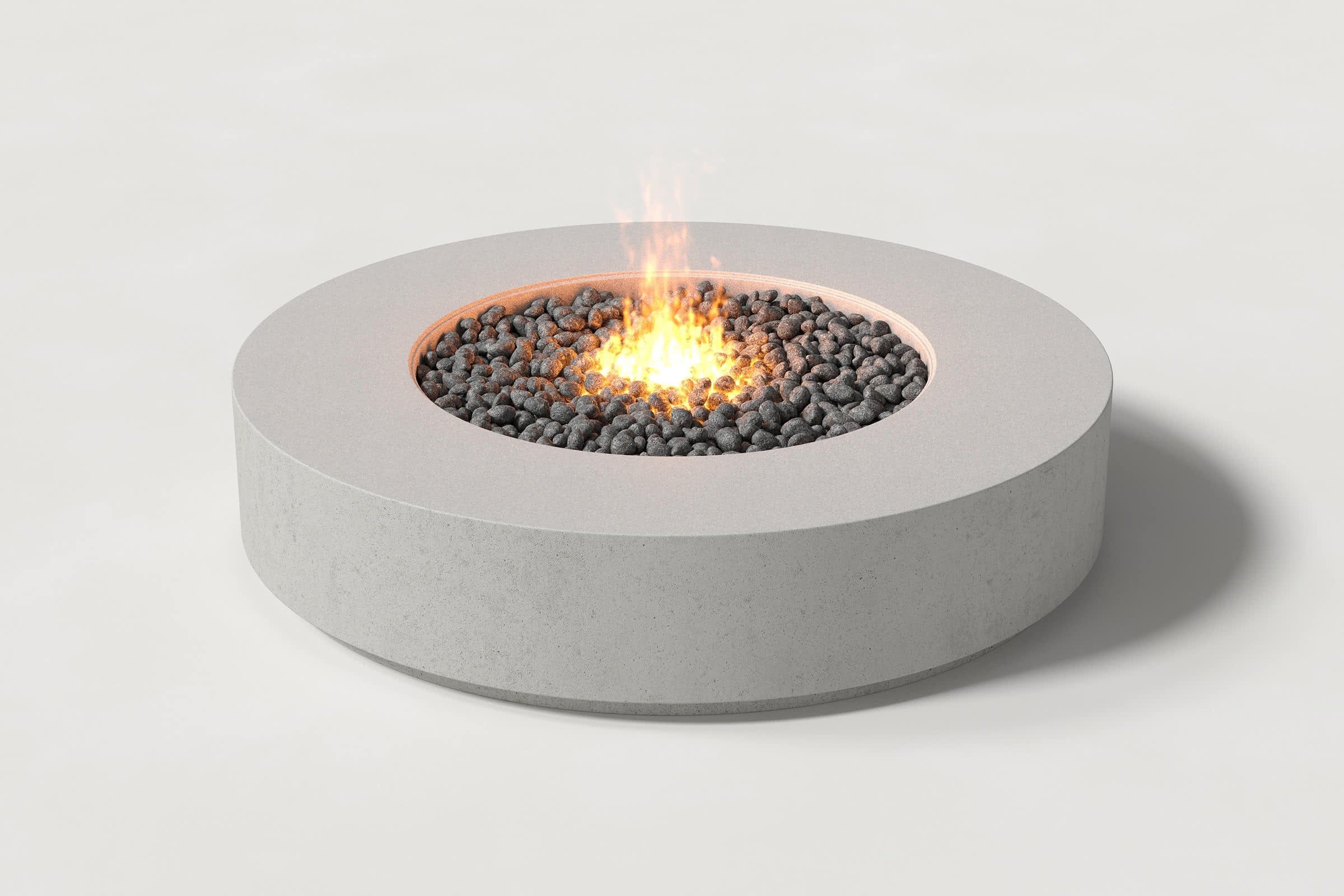 Low cylindrical soft grey concrete fire pit with thick walls and a central circular well. Mid-range angle shows the well filled with flaming volcanic rocks against a white background.