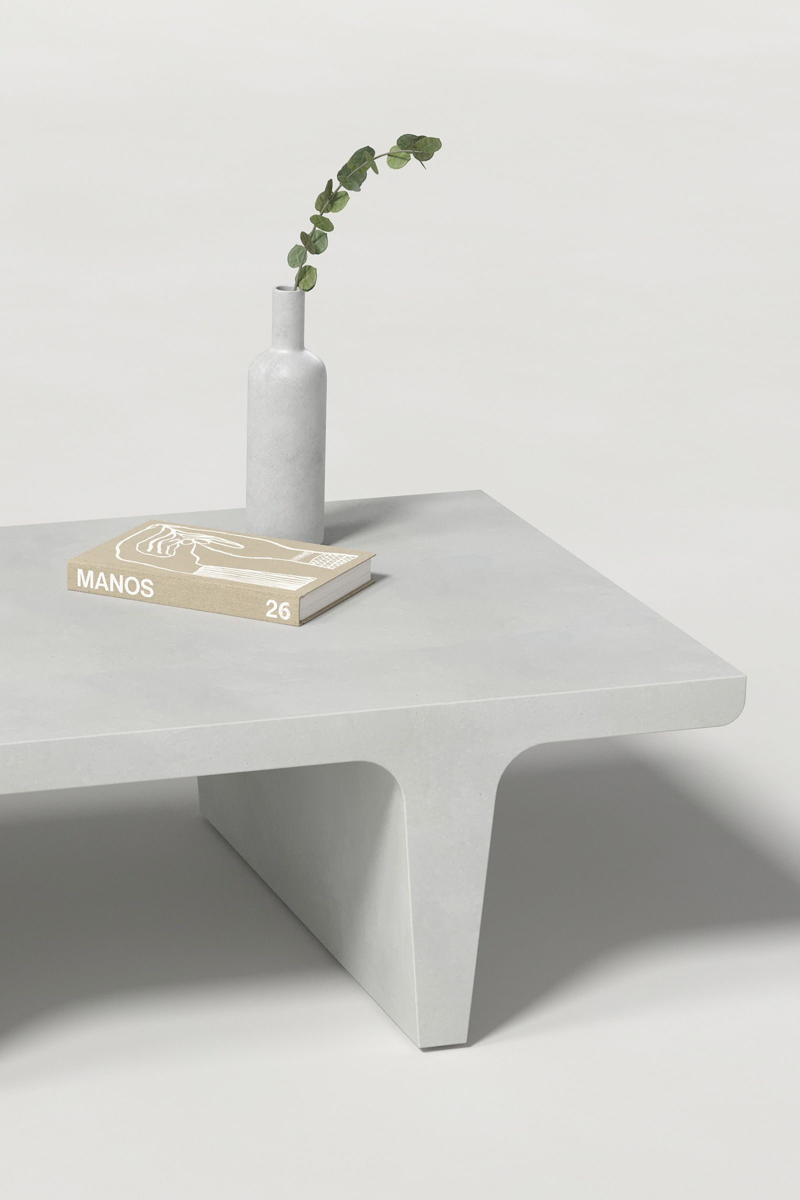 Close-up of a rectangular white concrete coffee table with sculptural double tee legs, styled with a neutral-toned book and a slender vase holding a eucalyptus, set against a clean white background.