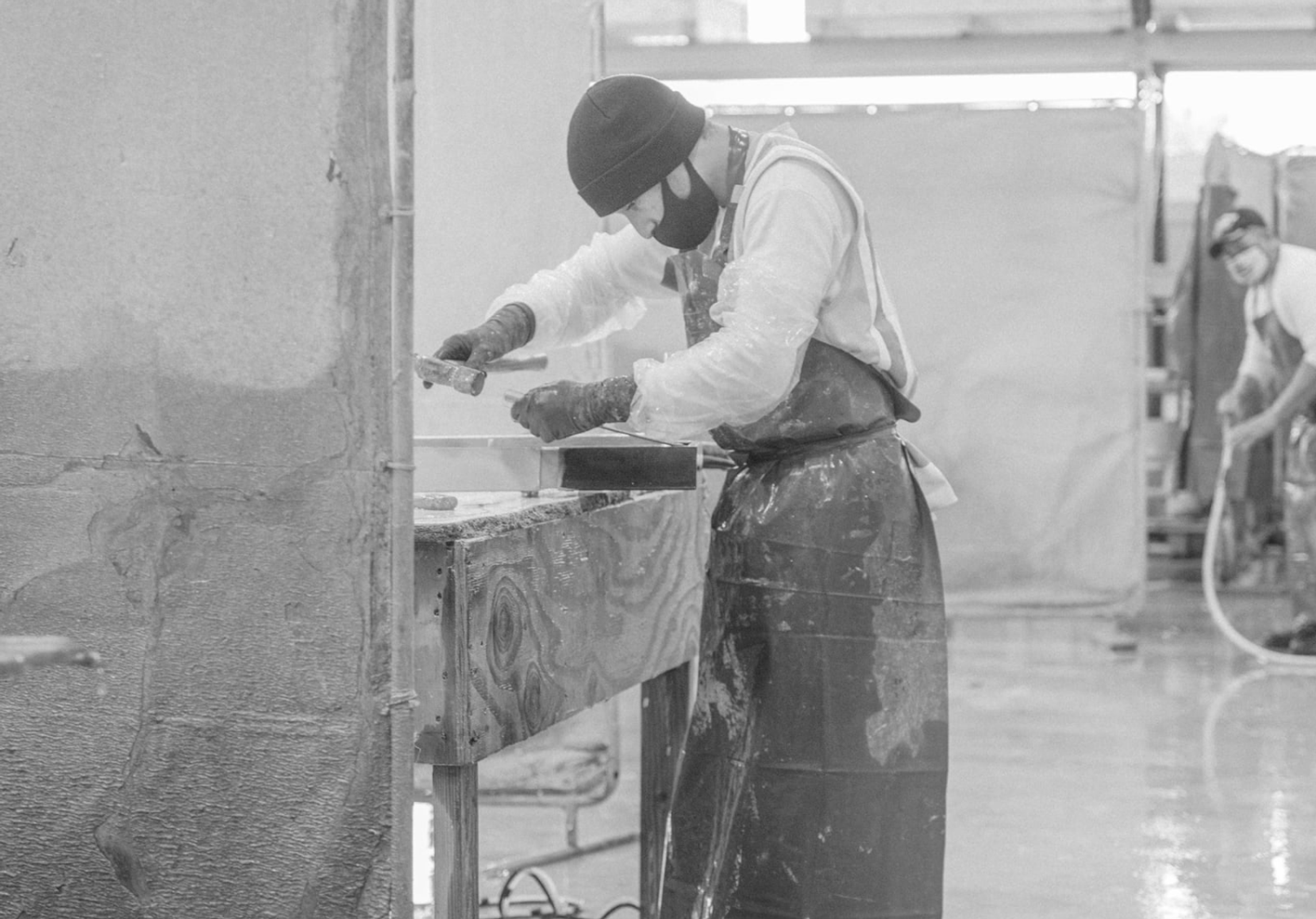 Concrete crafter in protective gear using a hammer to shape a concrete slab in a dusty worksite environment, shown in greyscale.