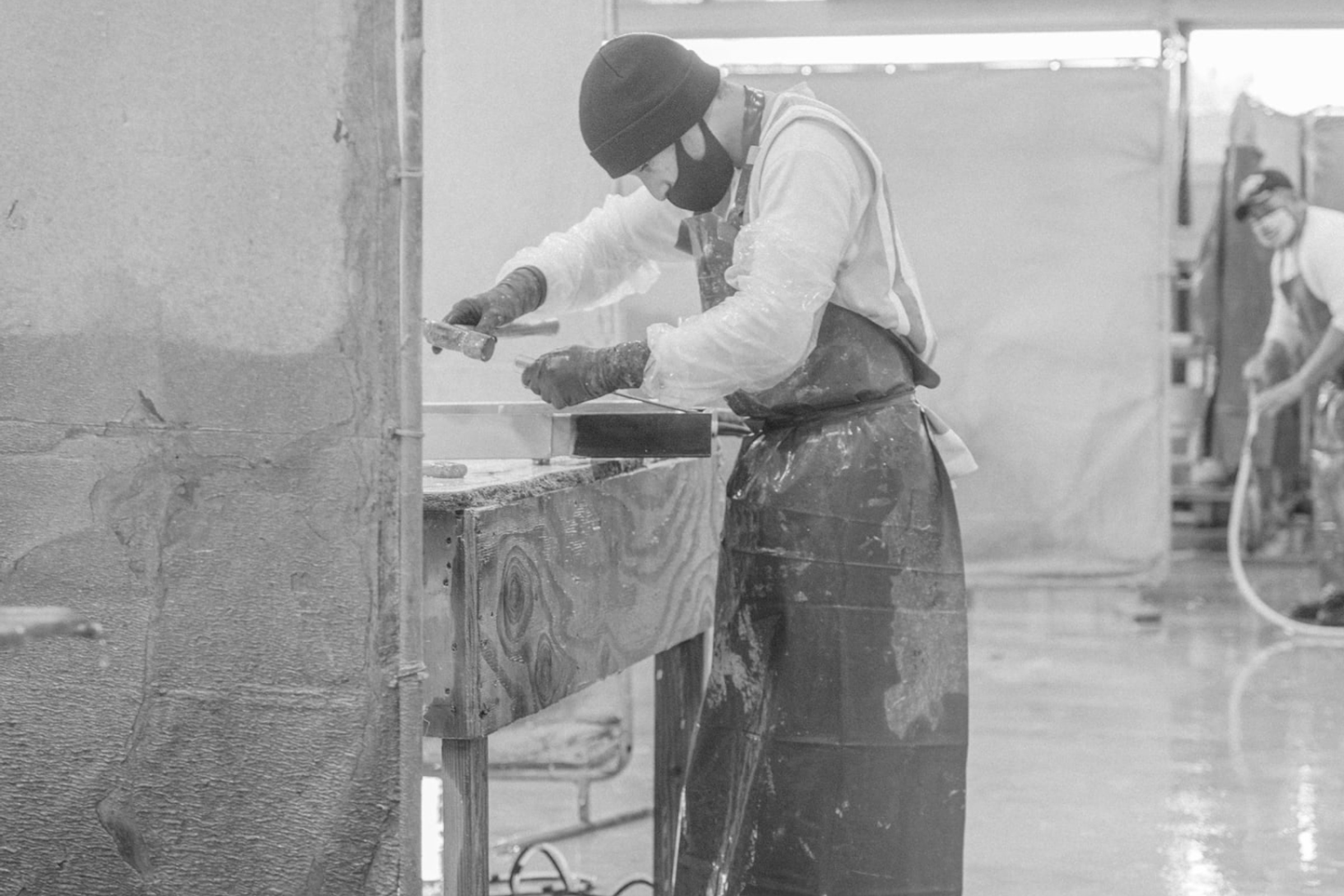 Concrete crafter in protective gear using a hammer to shape a concrete slab in a dusty worksite environment, shown in greyscale.