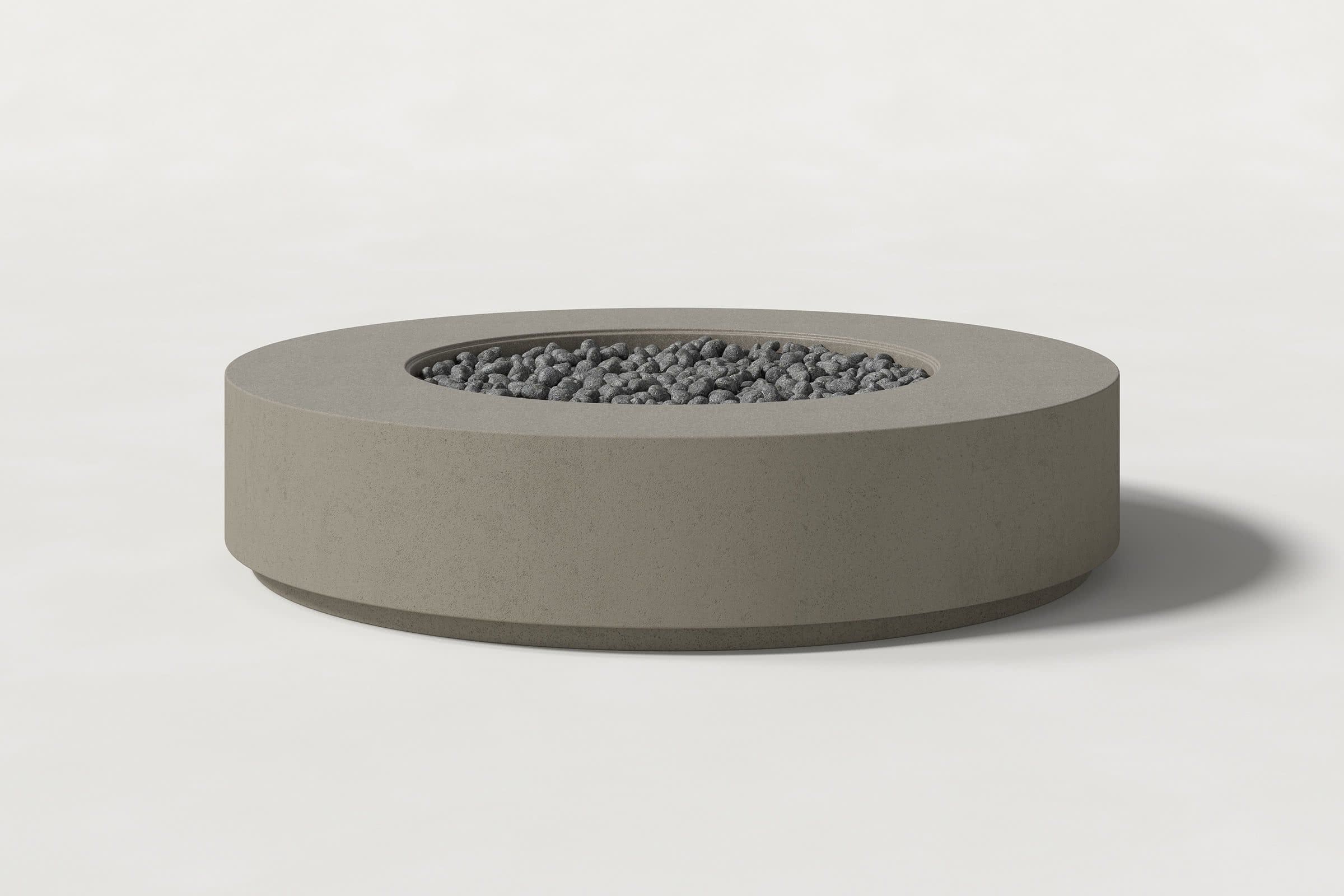 Low cylindrical dark grey concrete fire pit with thick walls and a central circular well filled with dark volcanic rocks. Side view on a white background with a shadow to the right.