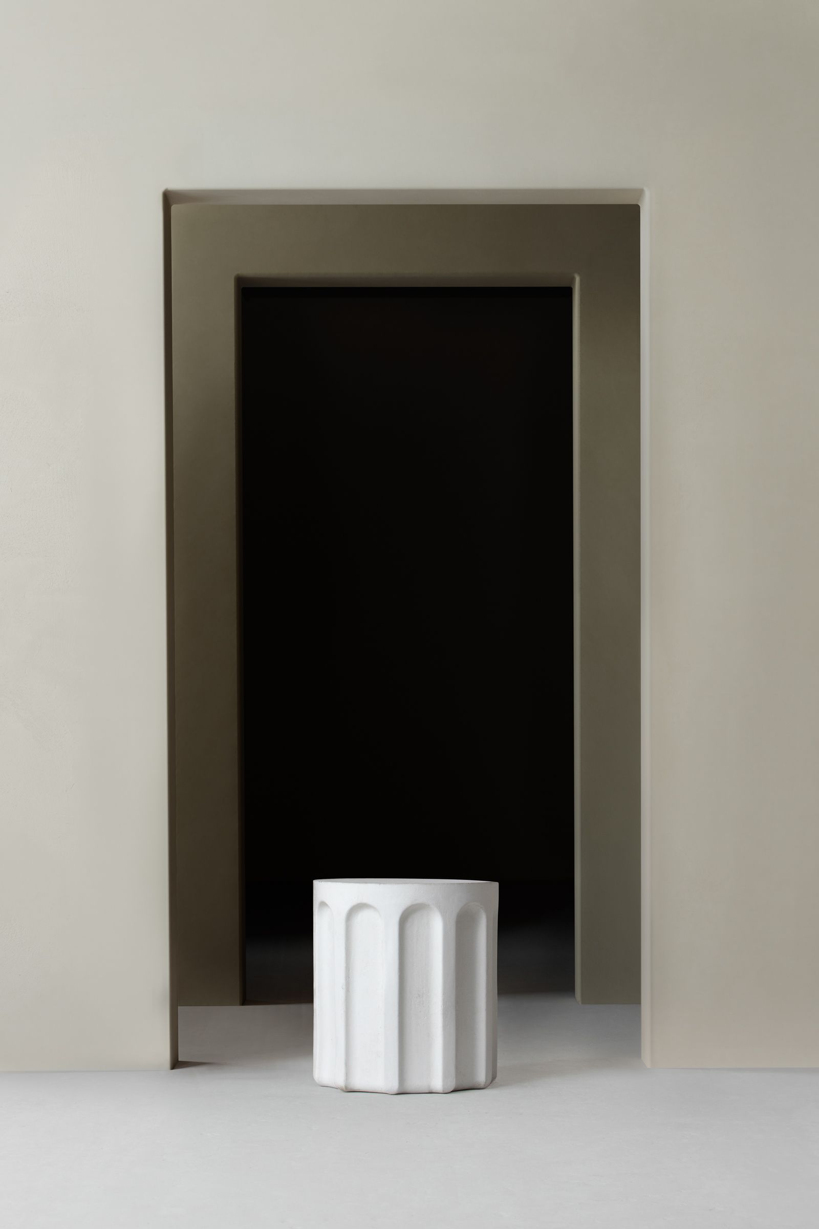 A sculptural white concrete side table with fluted detailing is centered in front of a large, dark recessed doorway. The surrounding walls and floor are smooth and neutral-toned, creating a minimalist and architectural setting.