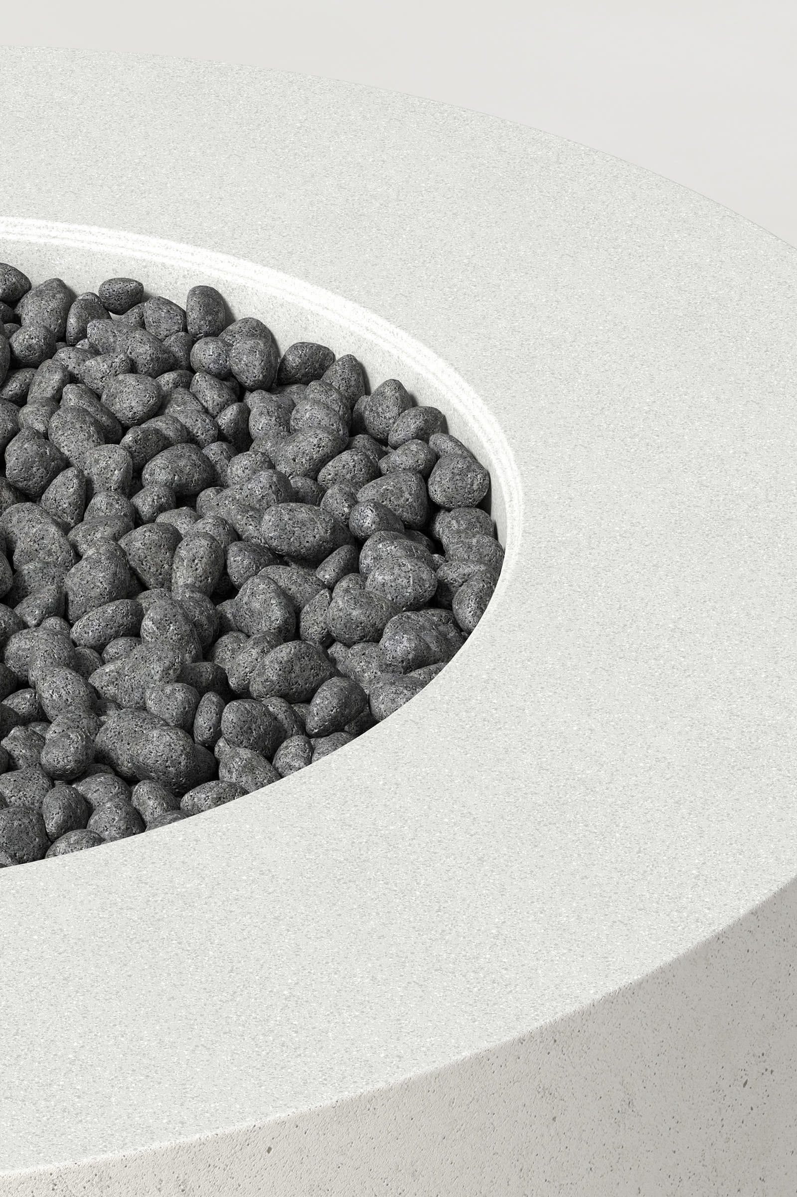Close-up photograph of a cylindrical white concrete fire pit with thick walls and a well filled with volcanic rocks. Showcases a smooth texture and precise edge design.