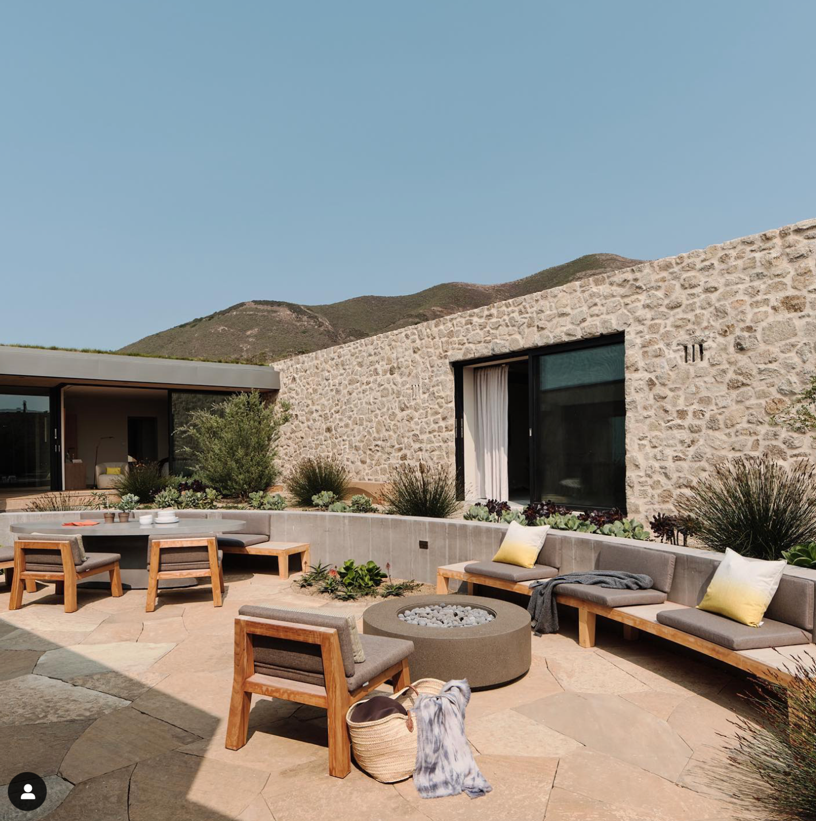 Modern stone-clad home with a sunlit courtyard featuring built-in bench seating, wood-framed lounge chairs, and a round concrete fire pit. Surrounded by desert landscaping and mountain views.