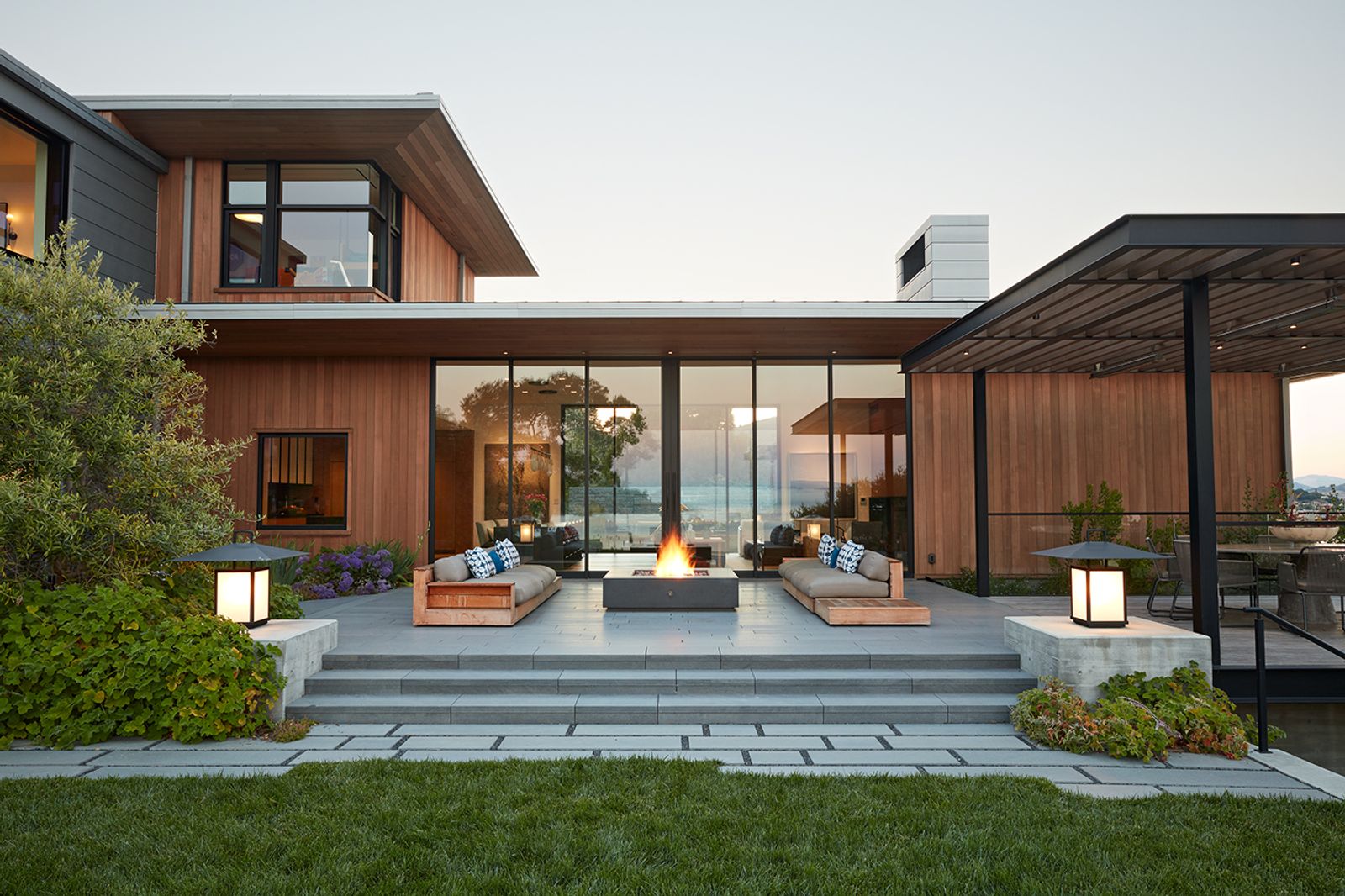 Modern cedar-clad home with floor-to-ceiling windows and outdoor living area featuring custom wood benches, large grey concrete fire pit, and  steps leading to a landscaped lawn.