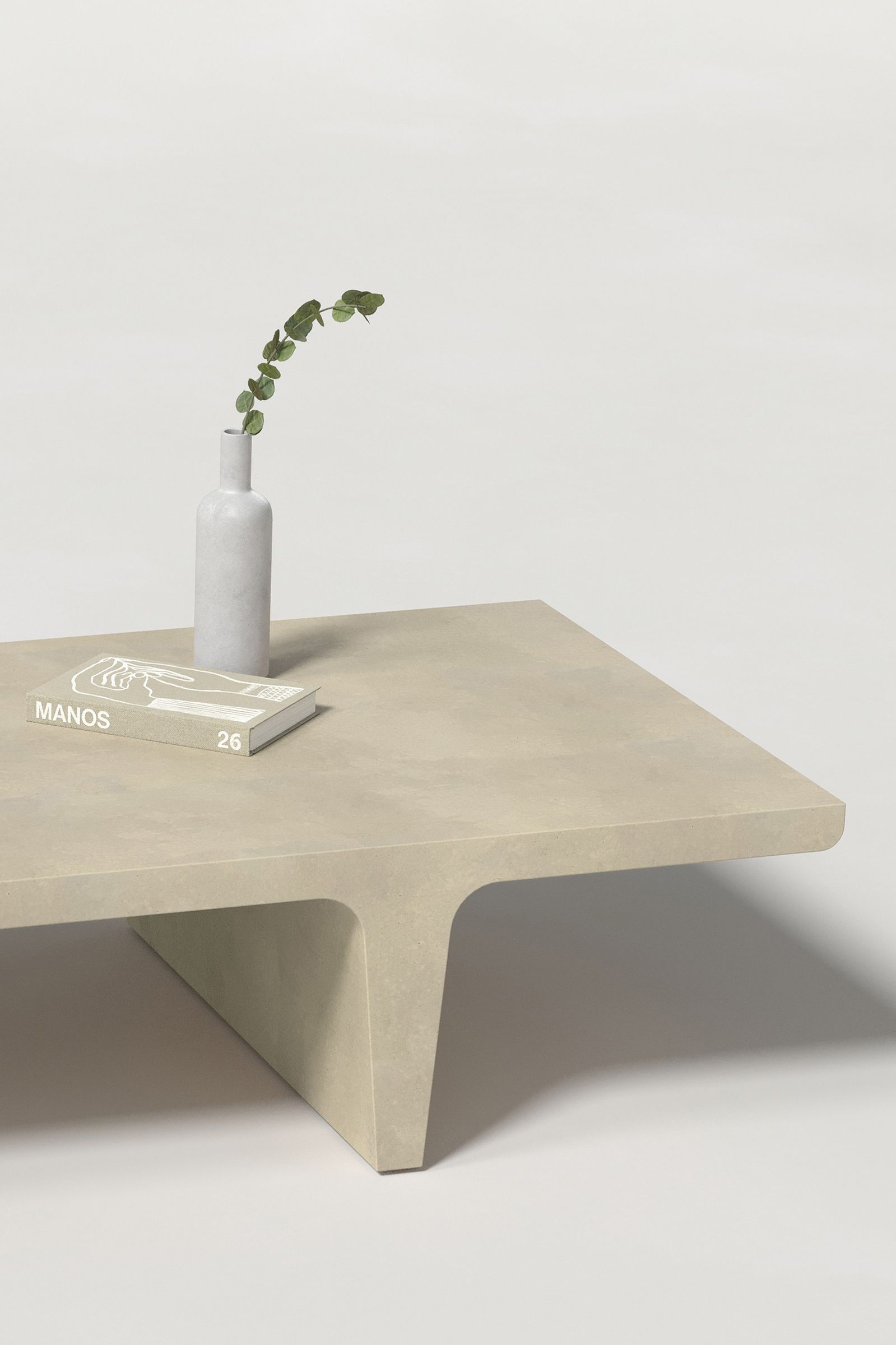 Close-up of a rectangular light beige concrete coffee table with sculptural double tee legs, styled with a neutral-toned book and a slender vase holding a eucalyptus, set against a clean white background.