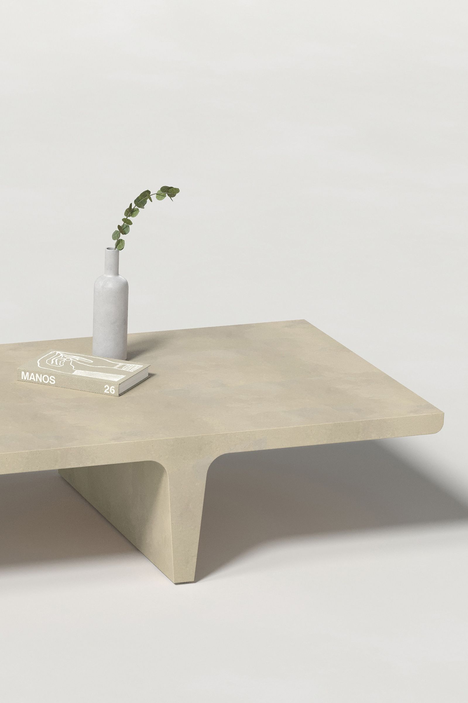 Close-up of a long rectangular light beige concrete coffee table with sculptural double tee legs, styled with a neutral-toned book and a slender vase holding a eucalyptus, set against a clean white background.