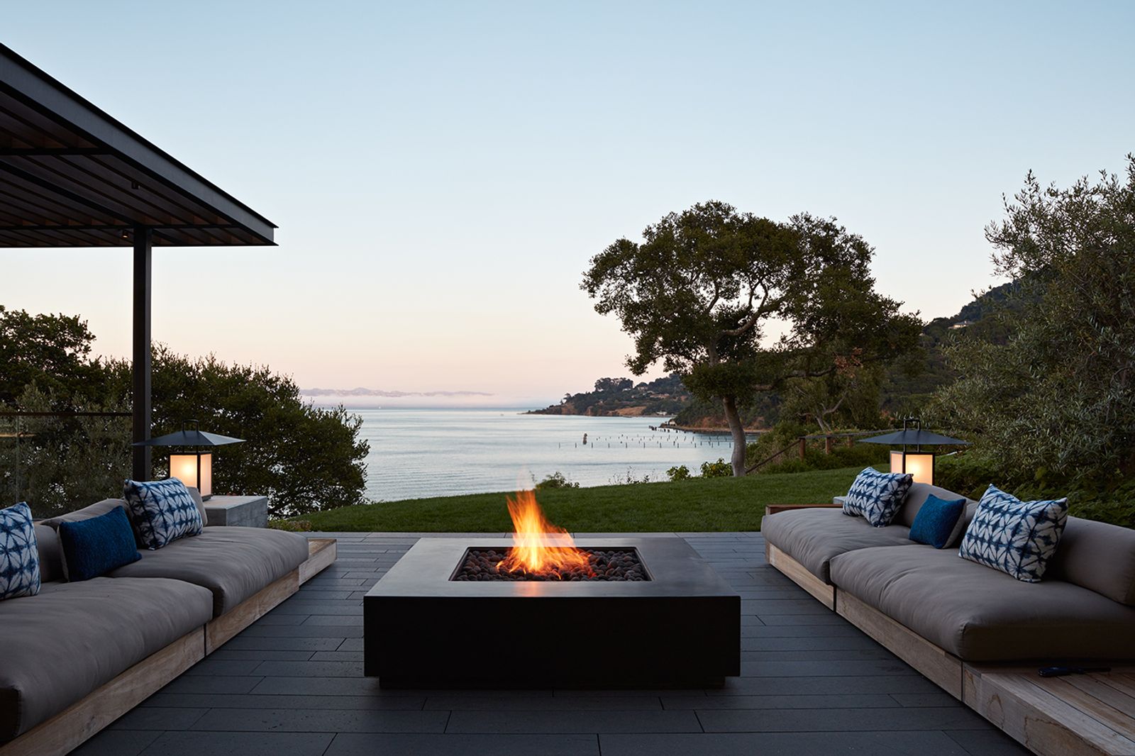 Contemporary waterfront patio with two outdoor sofas facing a dark grey, square concrete fire pit, overlooking a bay at sunset. Framed by trees and soft landscape lighting with a view of the ocean and hills.
