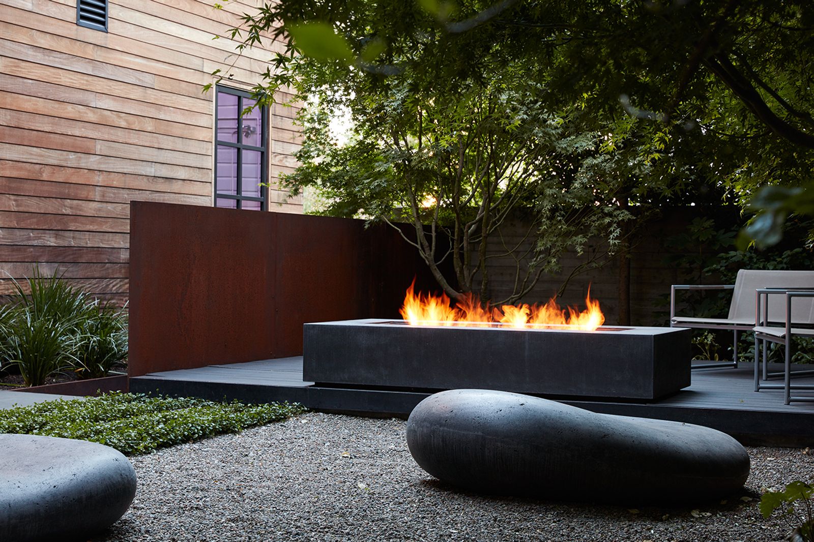 A backyard patio with a lit concrete fire pit and Soma stones. Backgrounded by a wooden house, trees and plants.
