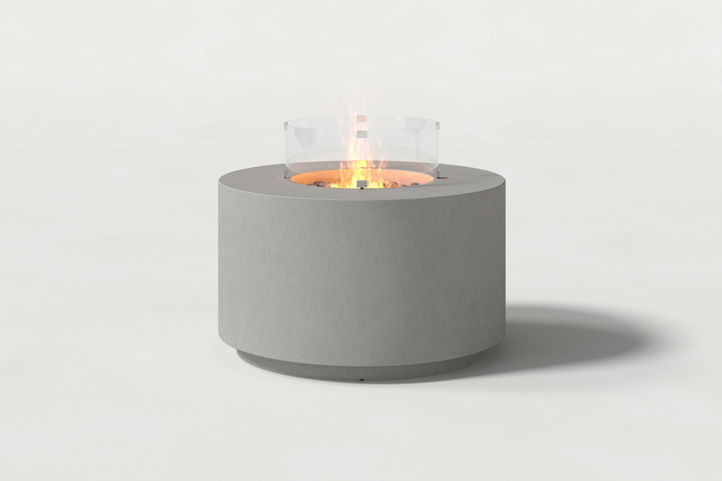 Low cylindrical grey concrete fire pit with thick walls and a central circular well filled with dark volcanic rocks and a glass shield. Side view on a white background with a shadow to the right.