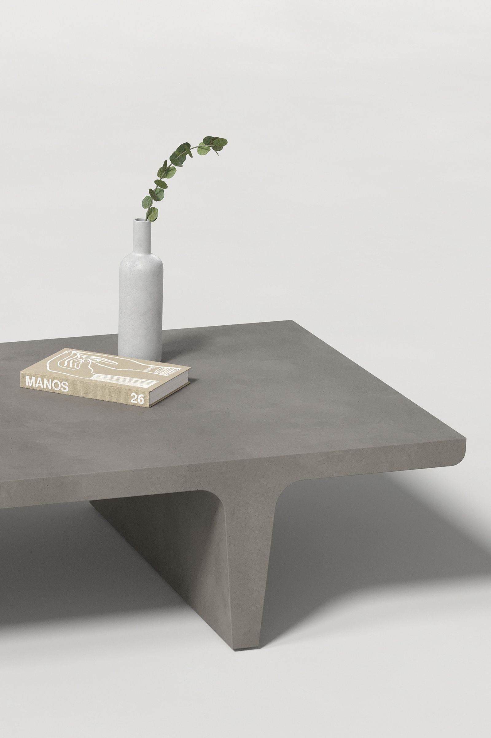 Close-up of a rectangular dark grey concrete coffee table with sculptural double tee legs, styled with a neutral-toned book and a slender vase holding a eucalyptus, set against a clean white background.