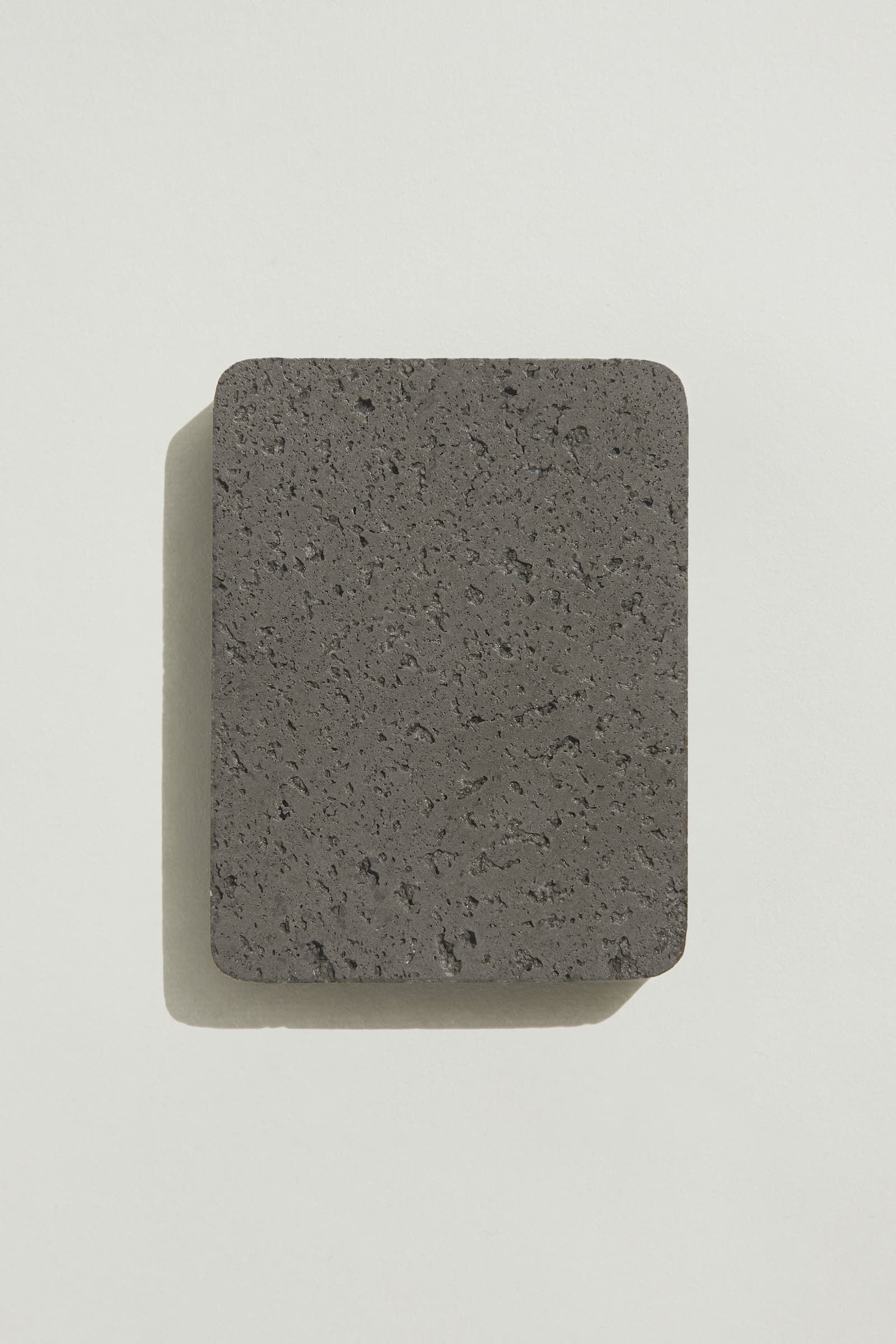 A rectangular sample of rough dark grey concrete with a natural, pitted surface texture. Photographed on a white background with a cast shadow.
