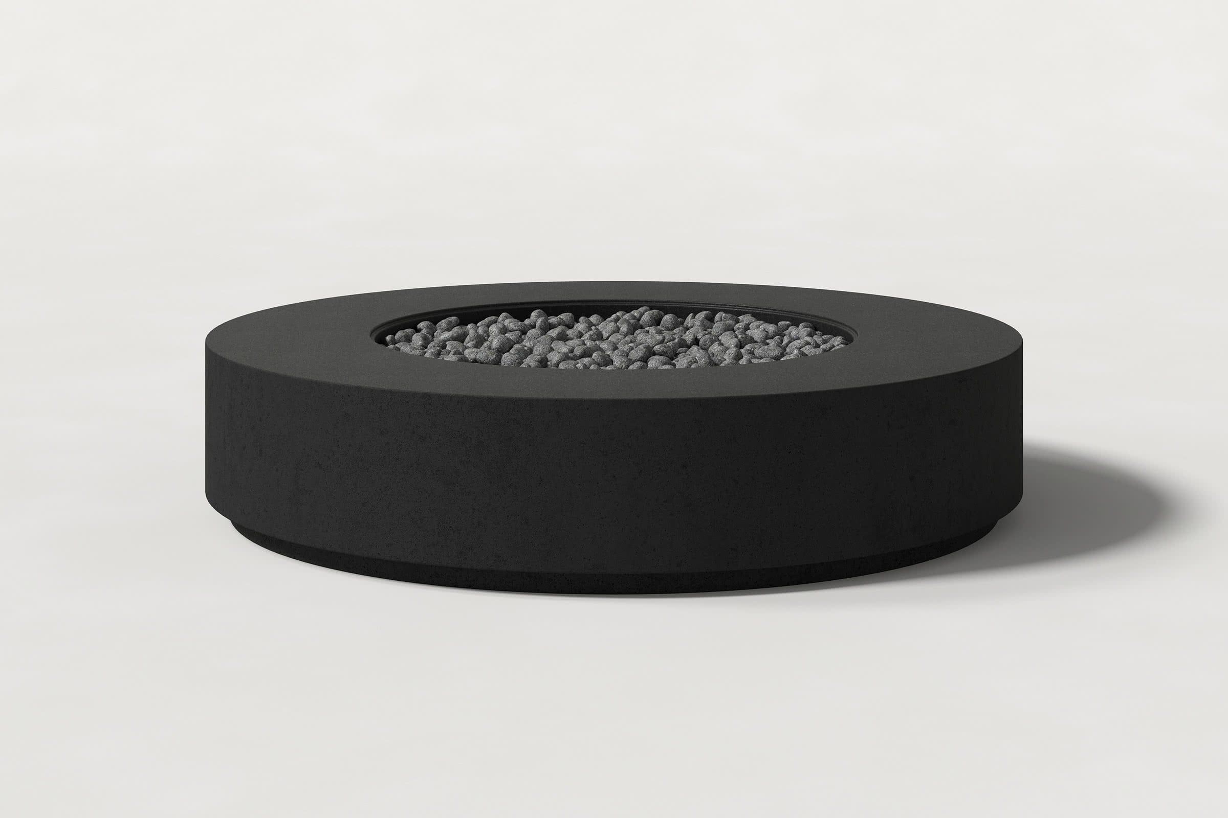 Low cylindrical jet black concrete fire pit with thick walls and a central circular well filled with dark volcanic rocks. Side view on a white background with a shadow to the right.