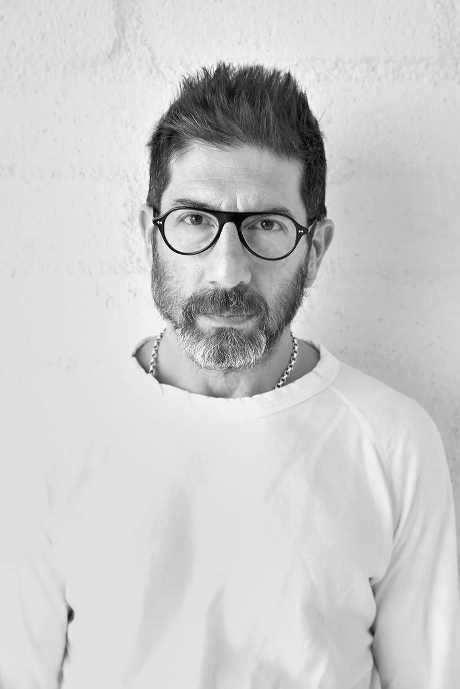 A black and white portrait photograph of a person wearing glasses and a white crew neck. They have short dark hair and beard. Standing against a plain light wall.