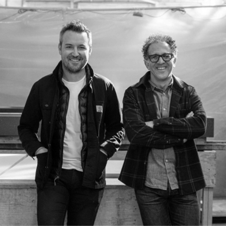 Two men standing side by side in a greyscale photo. Left man wears dark jacket over white shirt and hoodie, right man wears glasses and layered shirts with arms crossed.