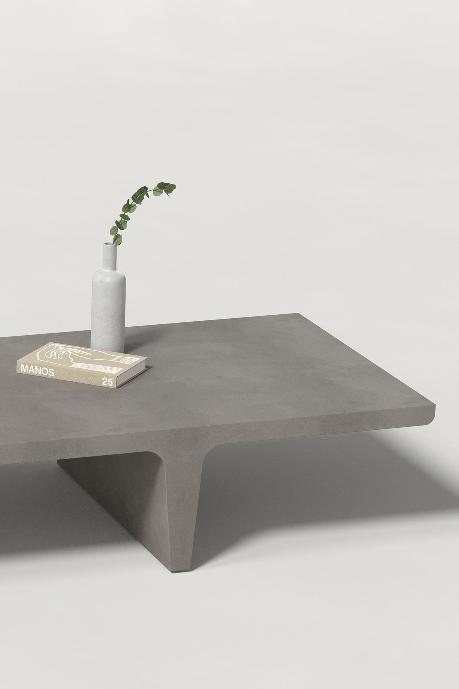 Close-up of a long rectangular dark grey concrete coffee table with sculptural double tee legs, styled with a neutral-toned book and a slender vase holding a eucalyptus, set against a clean white background.