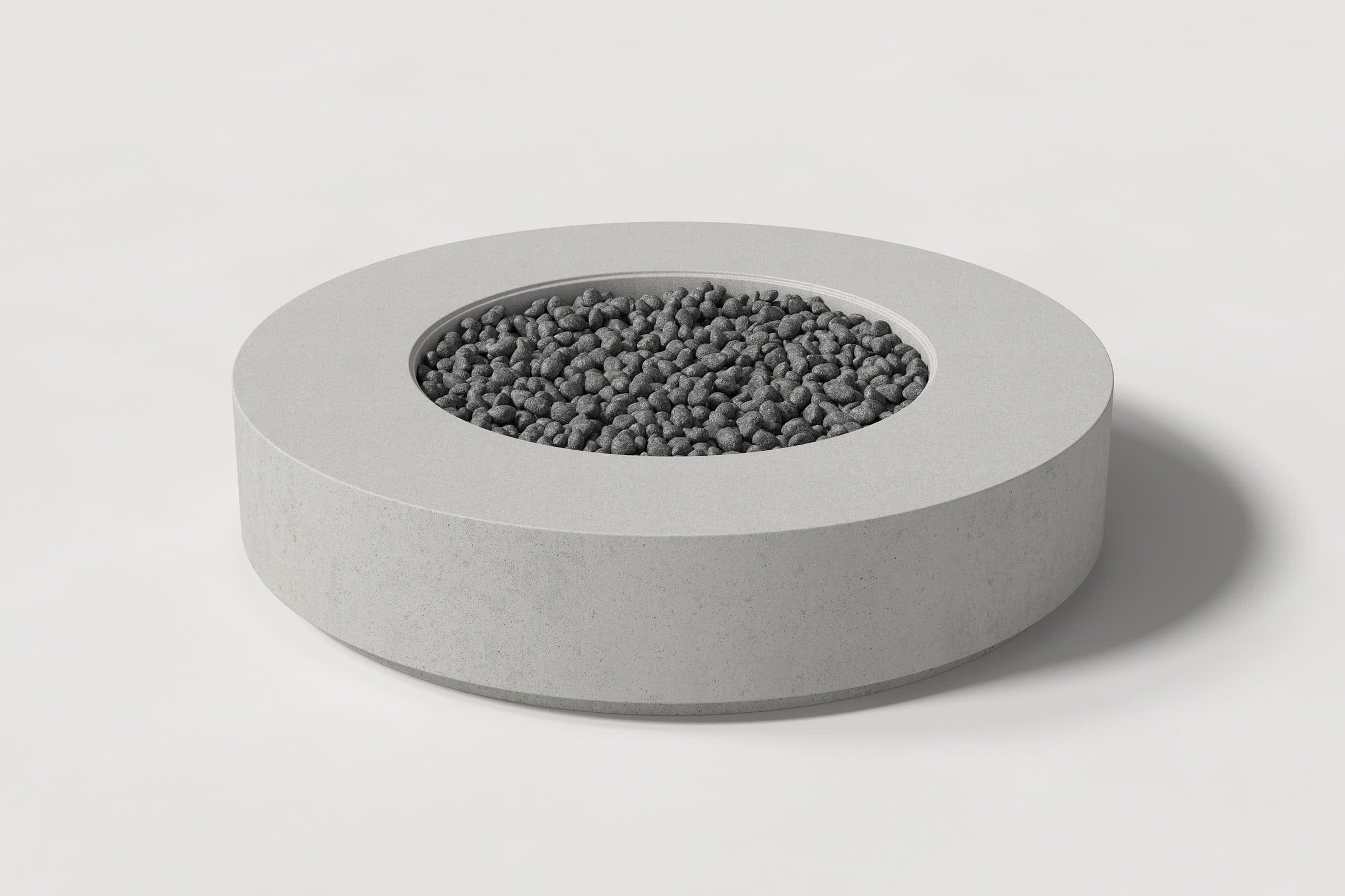 Low cylindrical soft grey concrete fire pit with thick walls and a central circular well. Mid-range angle shows the well filled with volcanic rocks against a white background.