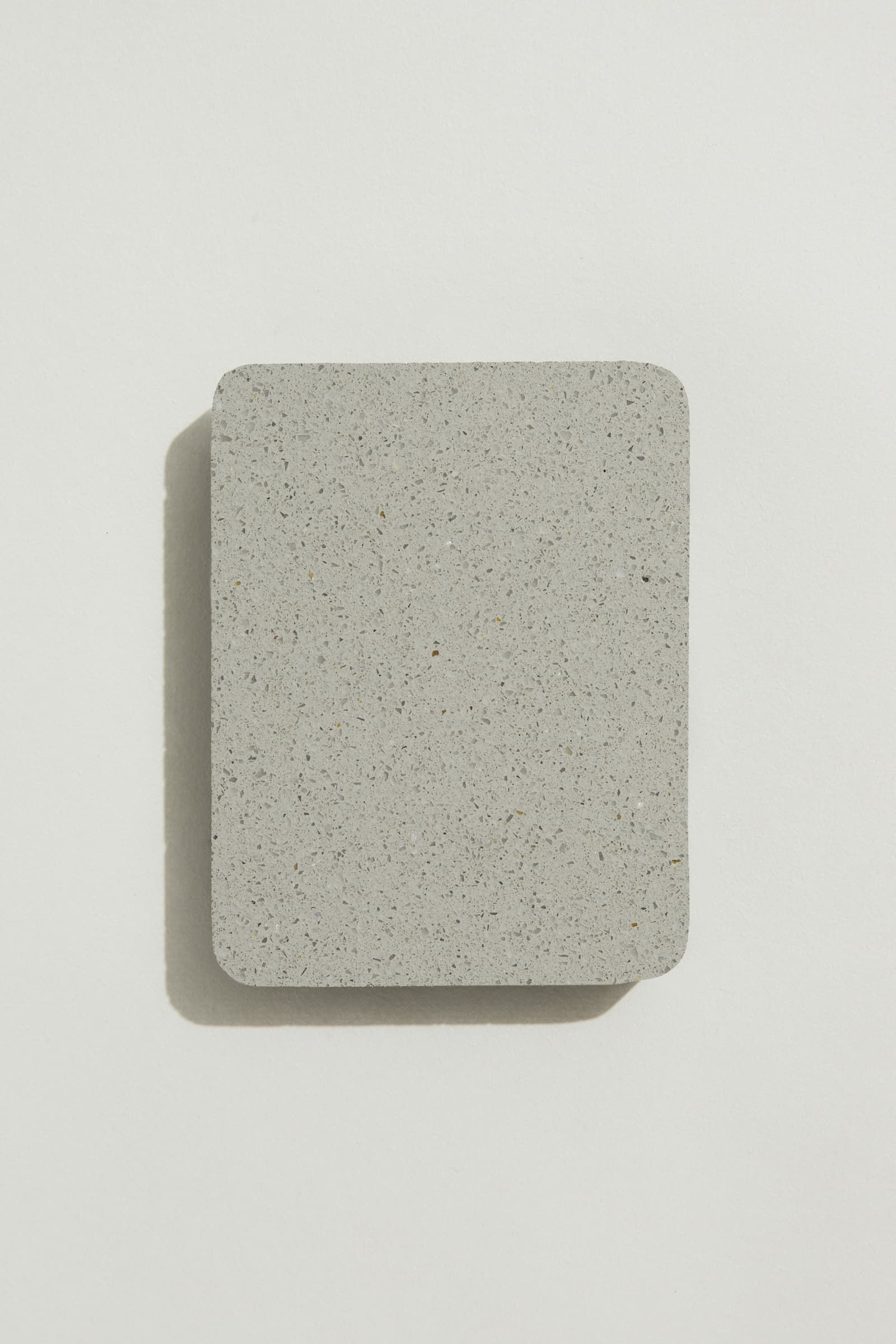 A rectangular sample of smooth soft grey concrete with grey, white and black terrazzo speckling, and rounded corners. Photographed on a white background with a cast shadow