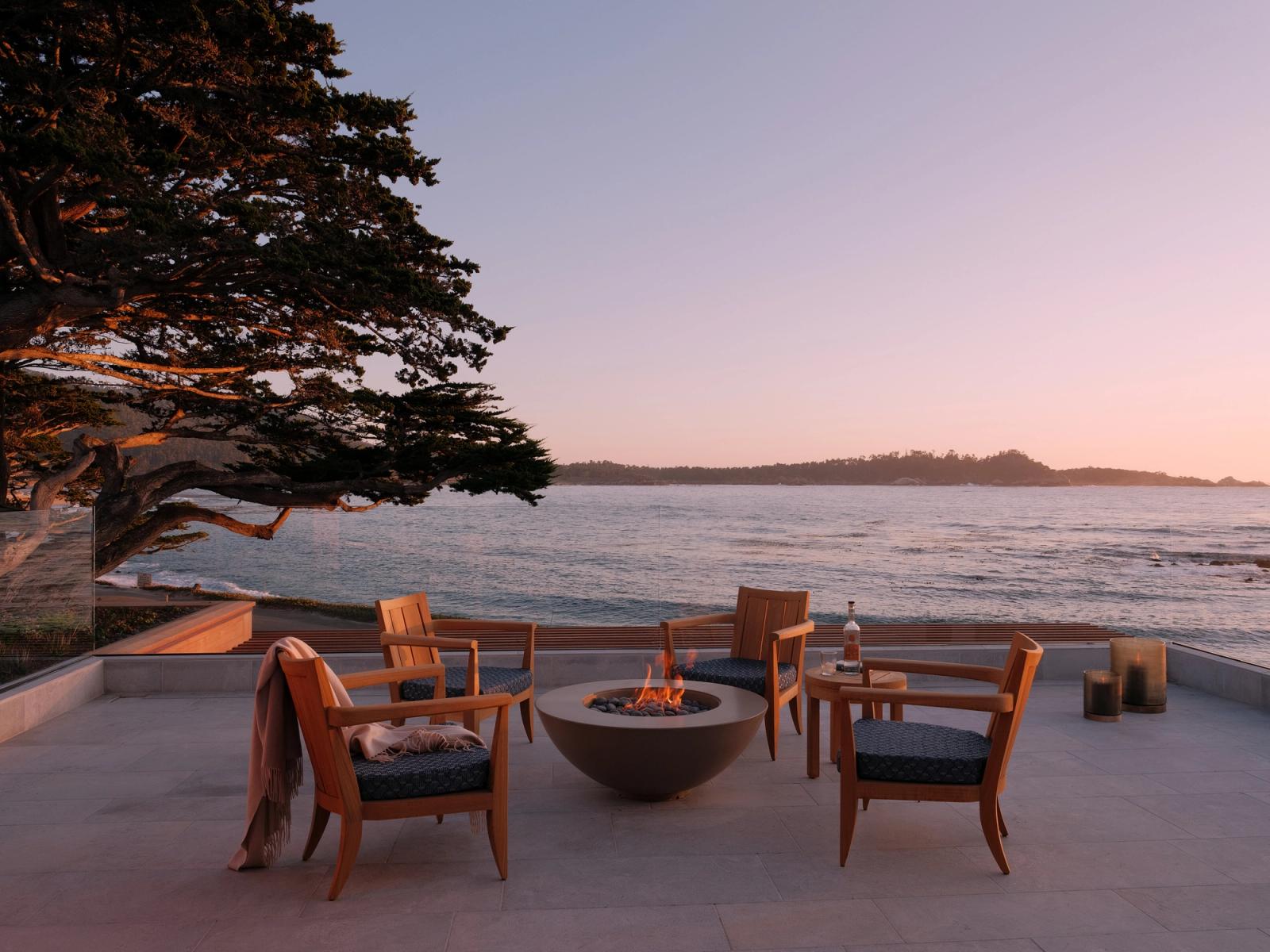 Oceanfront terrace at sunset with four wooden lounge chairs arranged around a round concrete lit fire pit. Overlooking calm coastal waters and distant shoreline, the space is framed by a windswept tree and soft evening light.