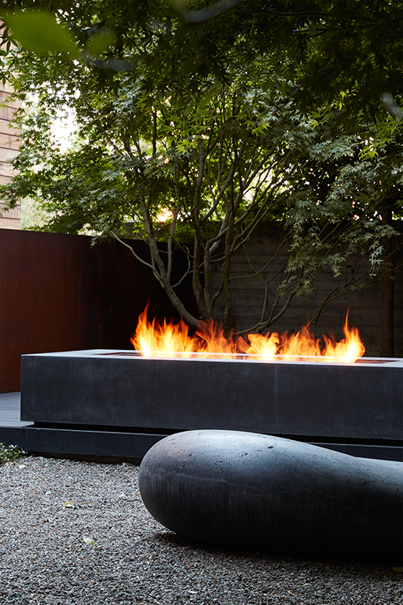 A backyard patio with a lit concrete fire pit and Soma stones. Backgrounded by a wooden house, trees and plants.