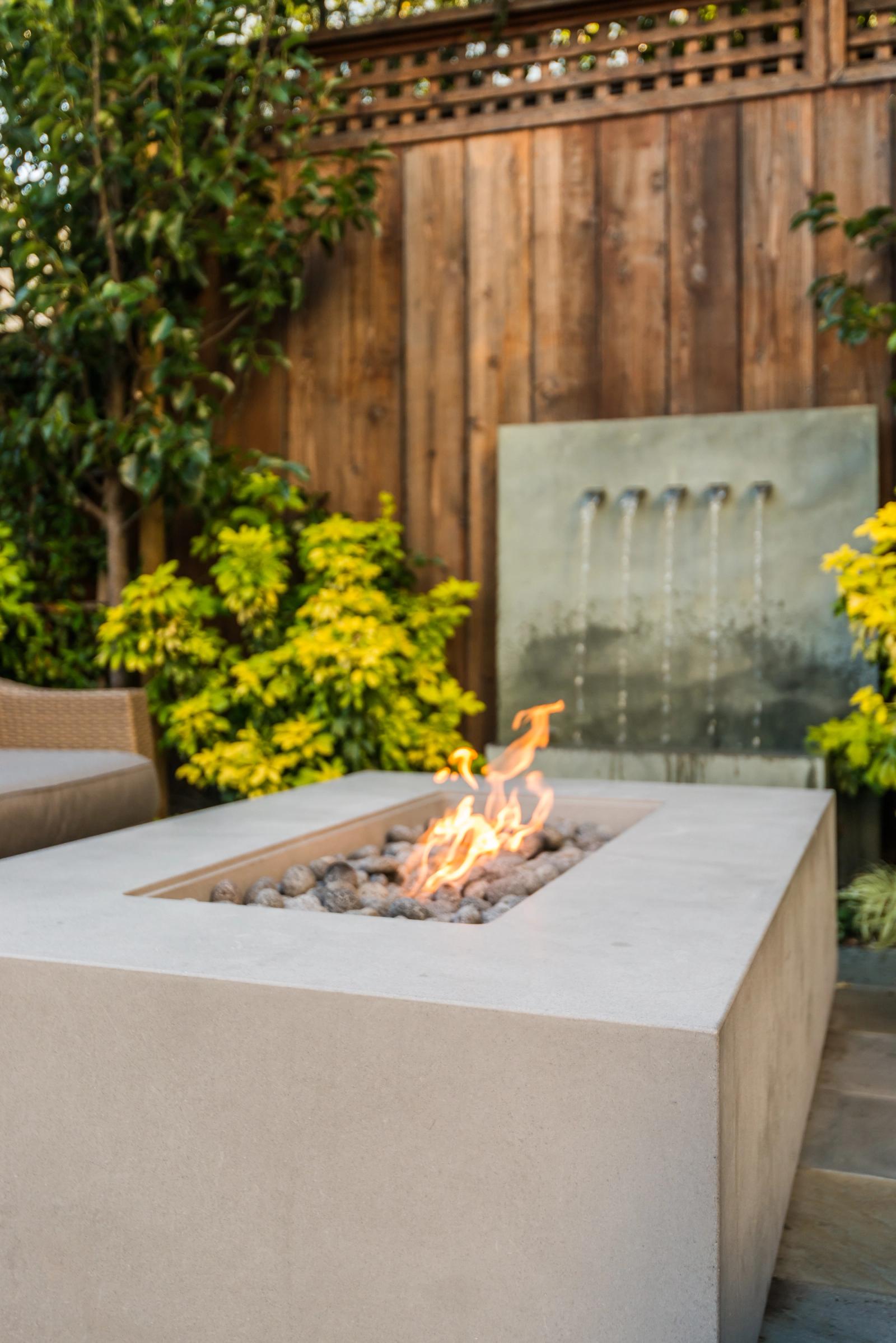 A backyard patio featuring a lit concrete fire pit table filled with stone and flames. Set against a wooden privacy fence, green and yellow foliage surrounds a modern wall water fountain into a basin behind it.