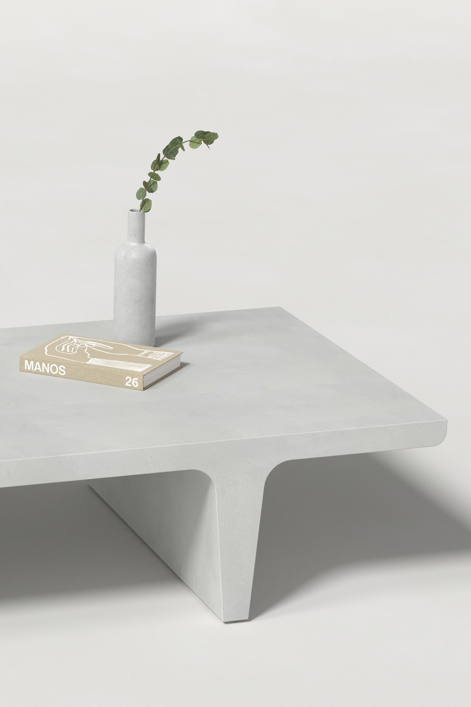 Close-up of a rectangular white concrete coffee table with sculptural double tee legs, styled with a neutral-toned book and a slender vase holding a eucalyptus, set against a clean white background.