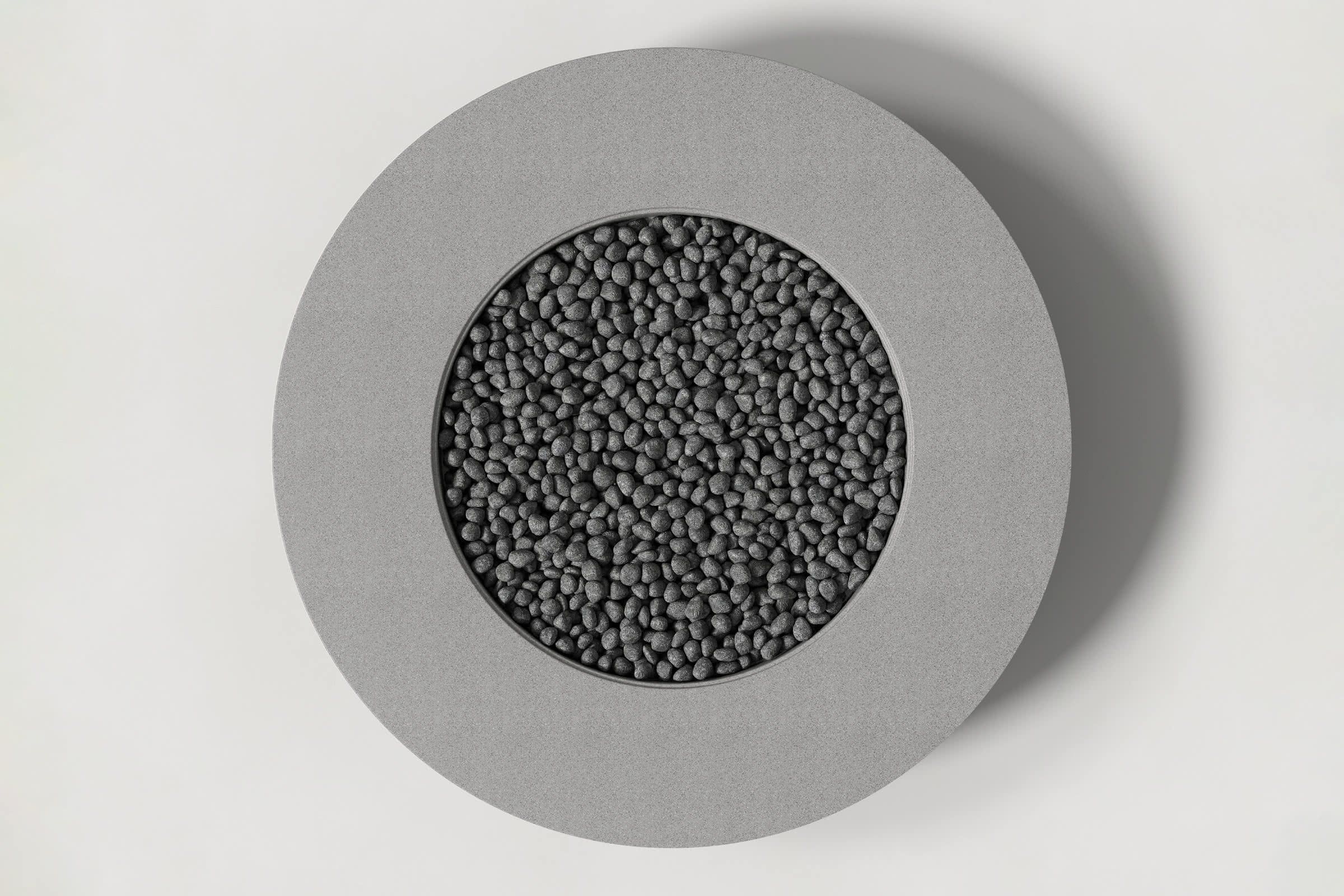 Top-down view of a circular grey concrete fire pit with thick walls and a circular well filled with volcanic rocks. Shown against a white background with slight shadows.