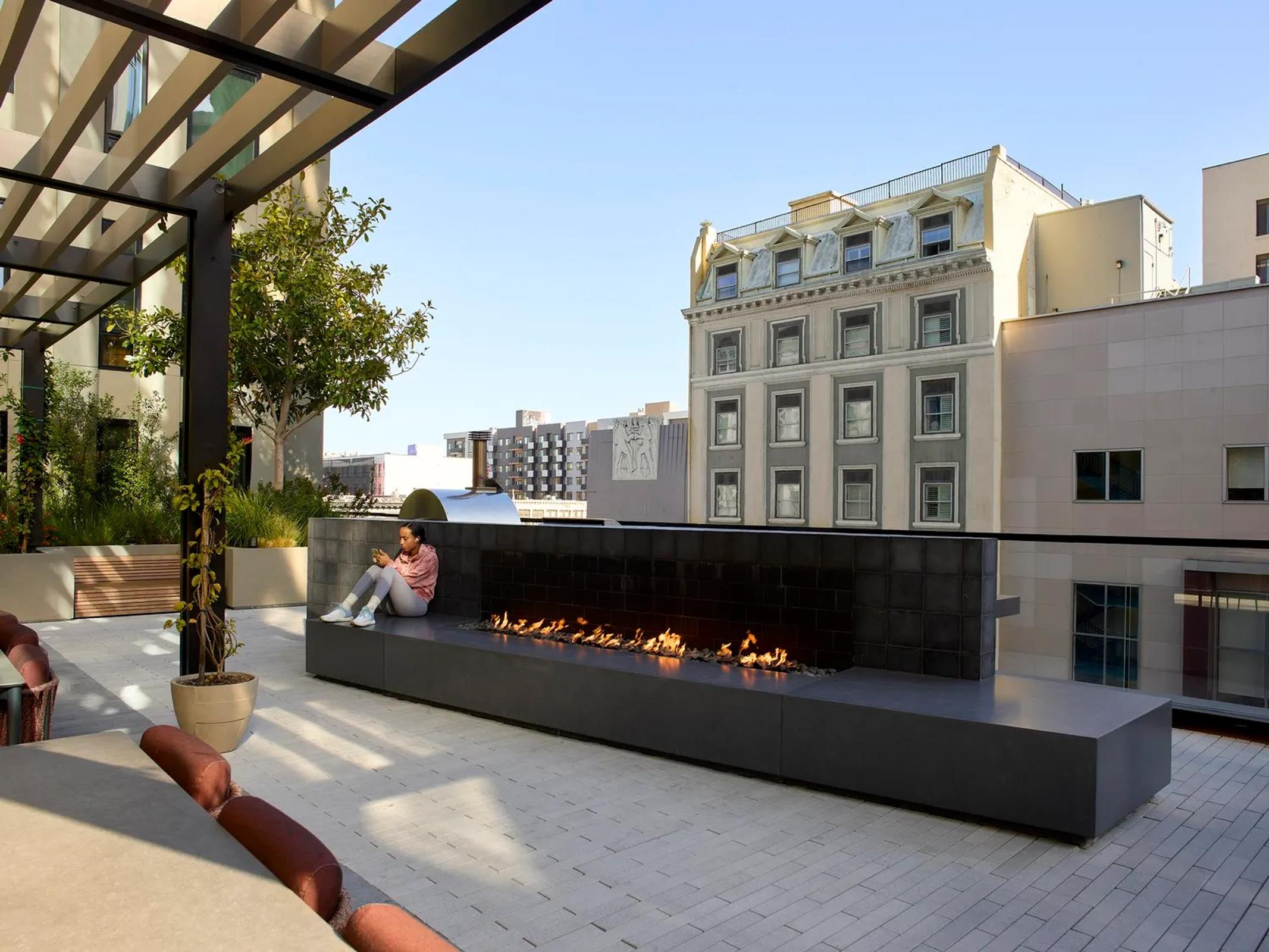 A modern rooftop terrace with long black fireplace. A person sits at its far edge. Includes a pergola, potted plants, leather seating, and views of the surrounding city