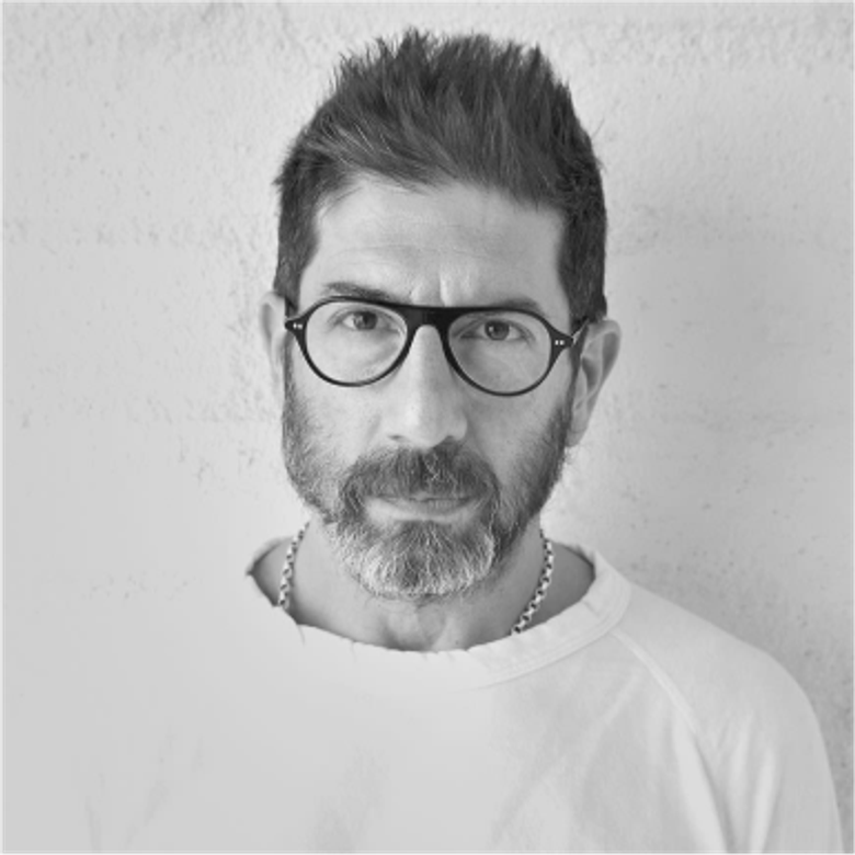 A black and white portrait photograph of a person wearing glasses and a white crew neck. They have short dark hair and beard. Standing against a plain light wall.