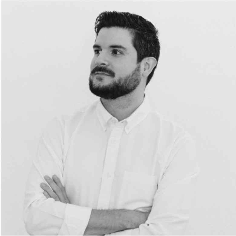 Black and white photo of a man with short dark hair and beard wearing a white button-up shirt, arms crossed, looking to the side against a light background.