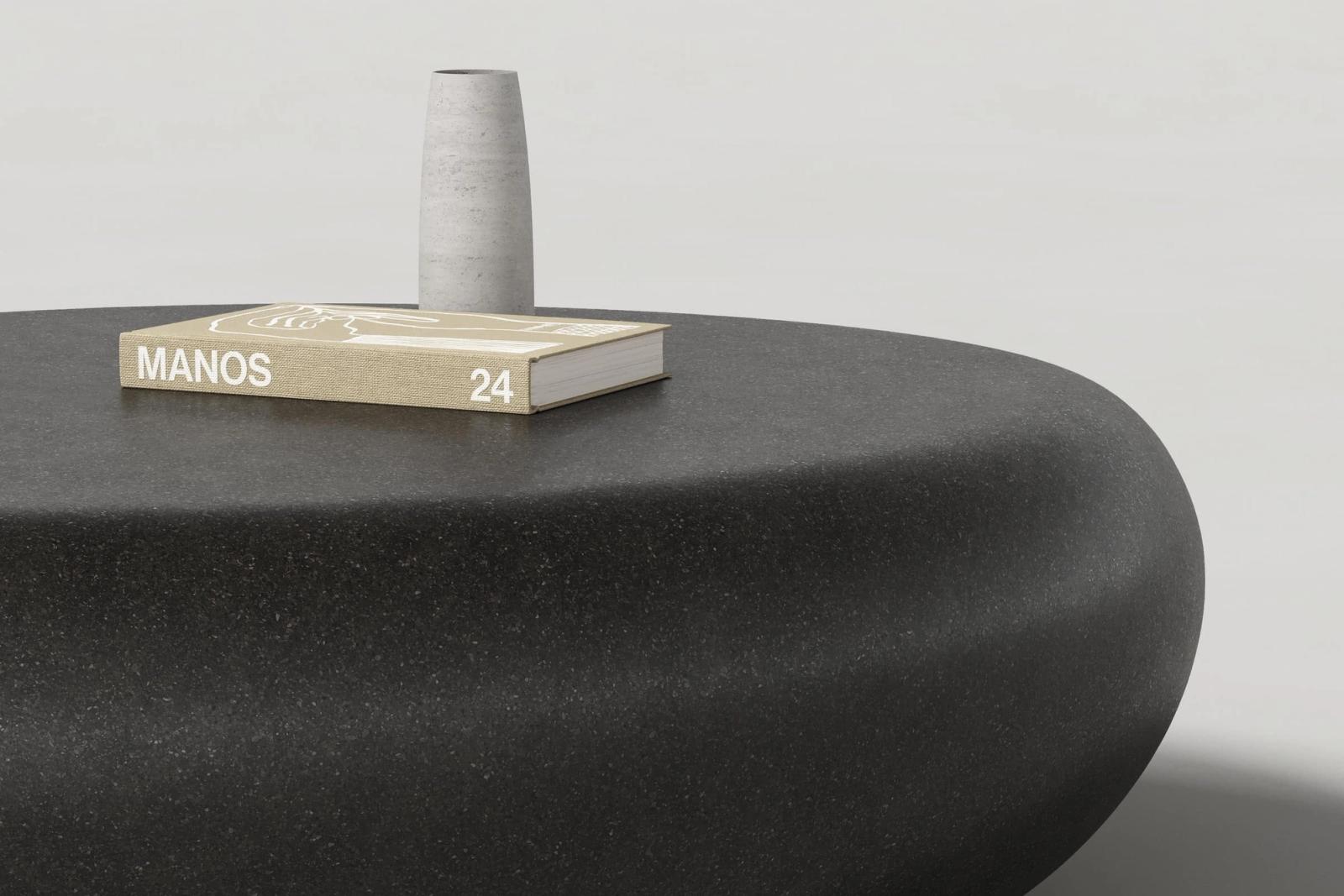 Close-up of a smooth jet black concrete coffee table with river rock-inspired form, smooth top and rounded edges. A beige MANOS 24 book and simple vase are displayed atop.