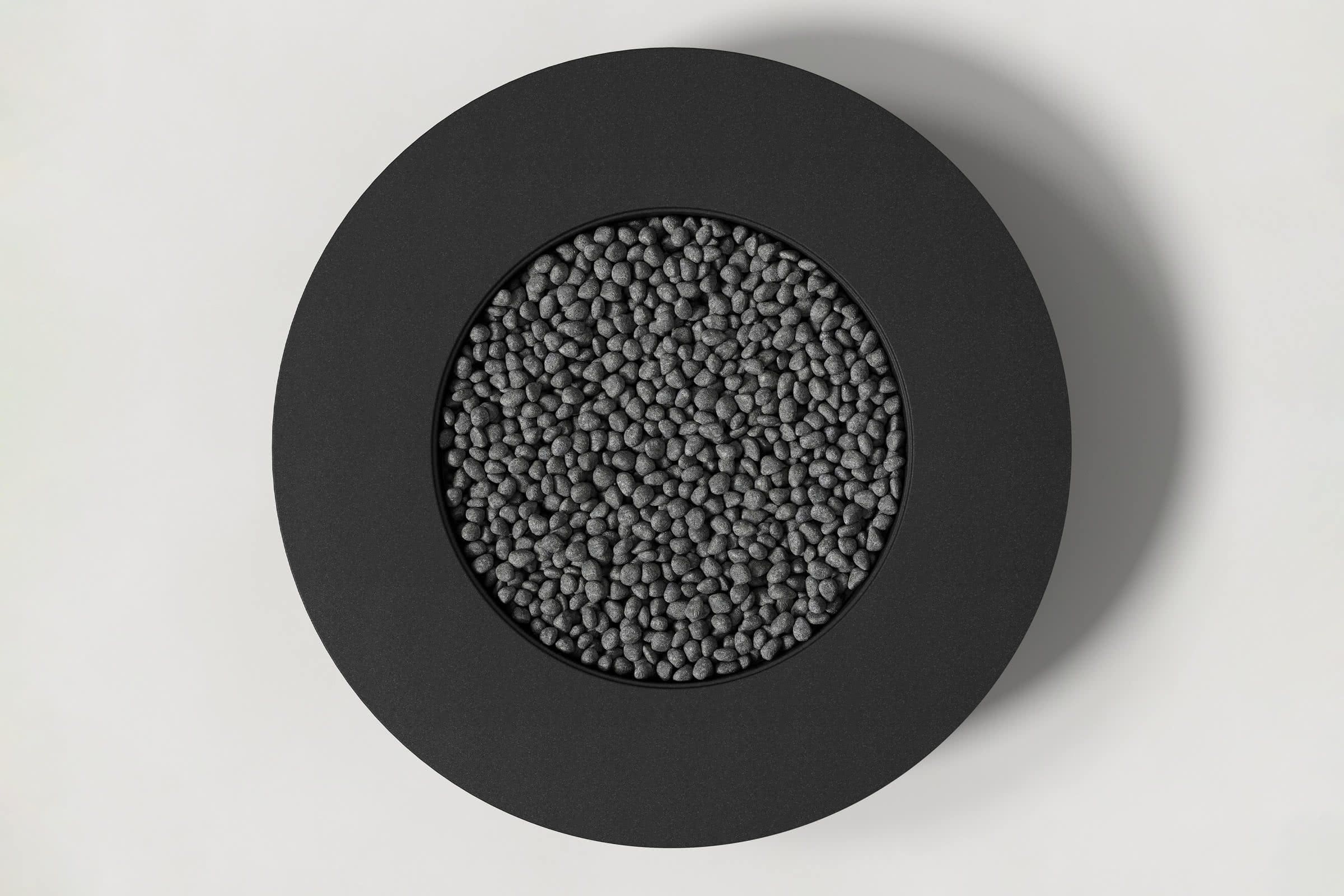 Top-down view of a circular jet black concrete fire pit with thick walls and a circular well filled with volcanic rocks. Shown against a white background with slight shadows.