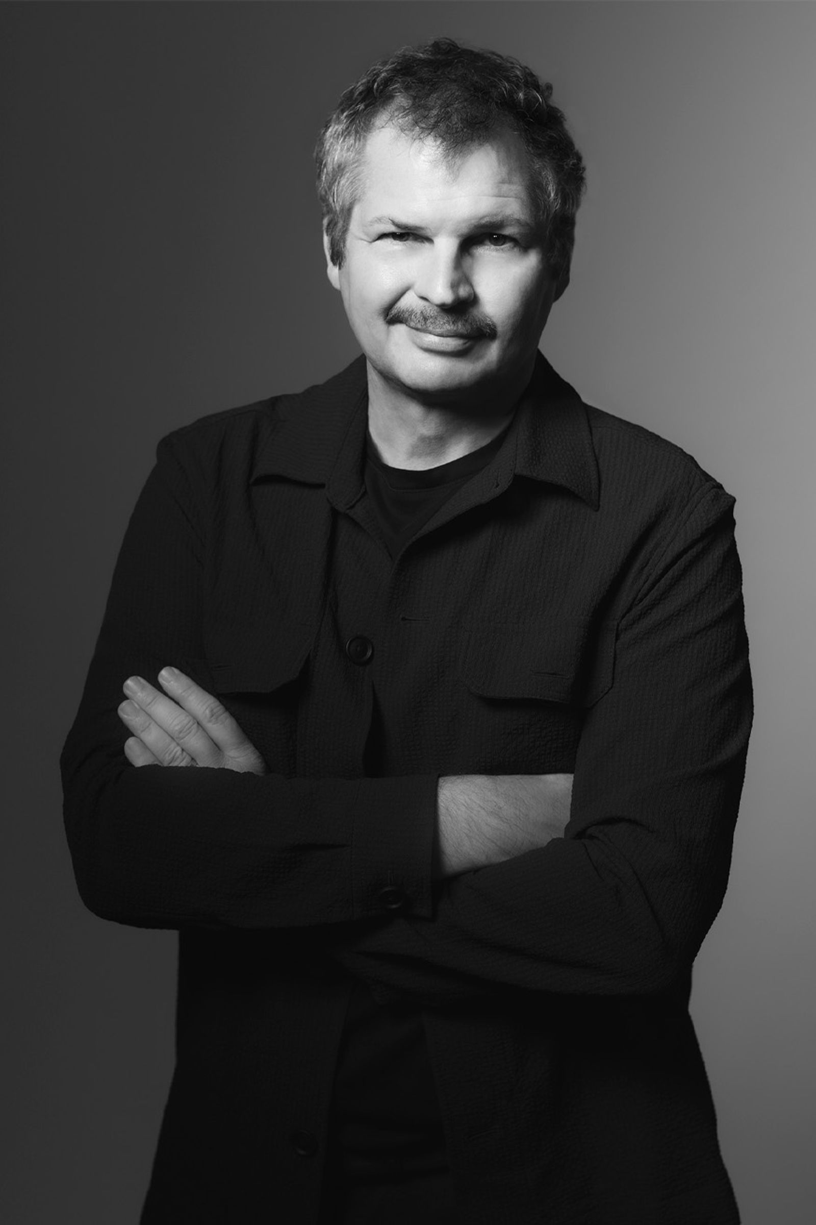 Black-and-white portrait of a man with short hair and a mustache, wearing a dark jacket and standing with arms crossed against a neutral gradient background. Max Strang.