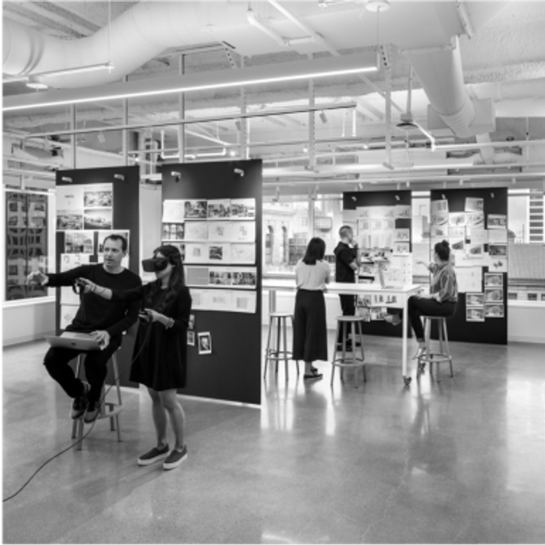 Black and white photo of an open office or design studio with several people working. Two people stand by display boards while others work at high tables.