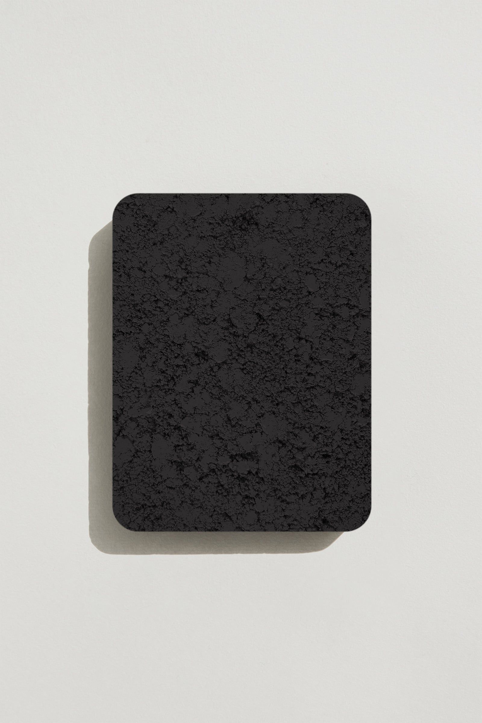 A rectangular sample of rough jet black concrete with a natural, pitted surface texture. Photographed on a white background with a cast shadow.