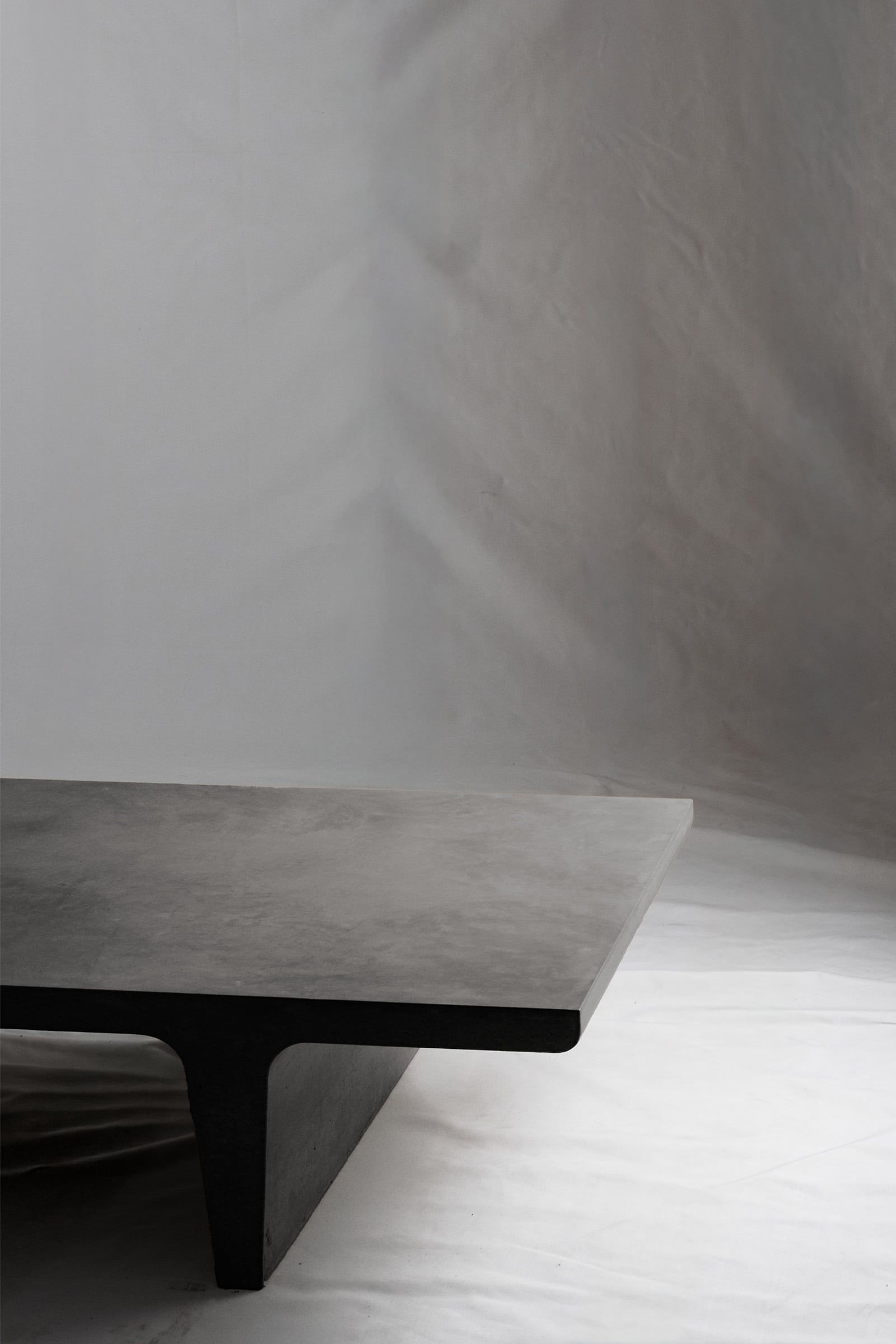 A close up of a dark grey concrete coffee table with sculptural double tee legs, featuring a smooth troweled finish, set against a soft neutral backdrop.