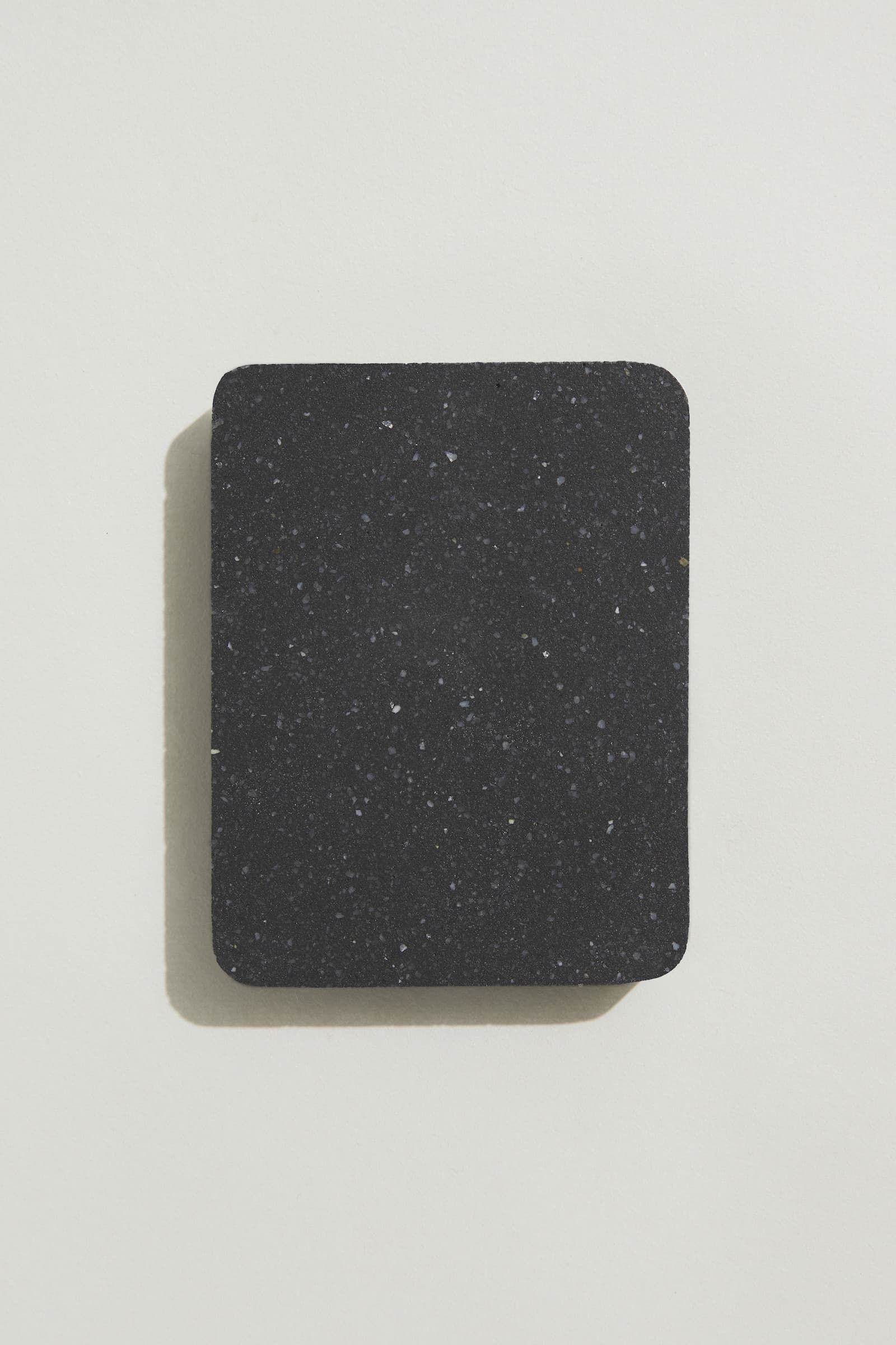 A rectangular sample of smooth jet black concrete with grey and white terrazzo speckling, and rounded corners. Photographed on a white background with a cast shadow