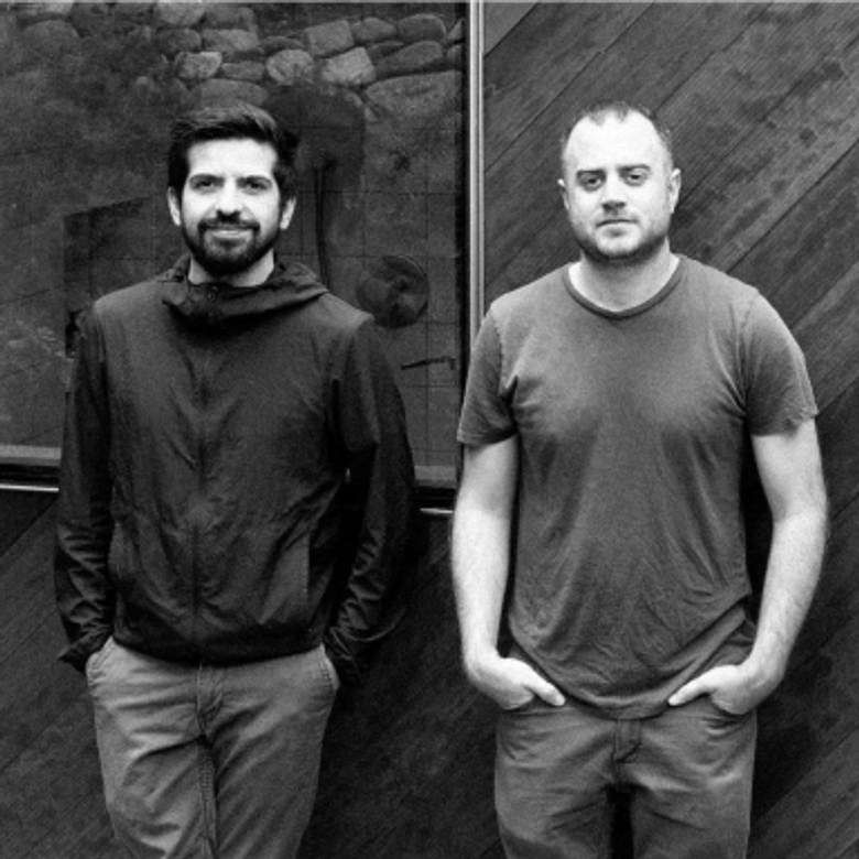 A black and white photograph of two people standing against dark contrasting stone & wooden wooden background. Both are wearing casual attire, one in a long-sleeved shirt , the other in a t-shirt.