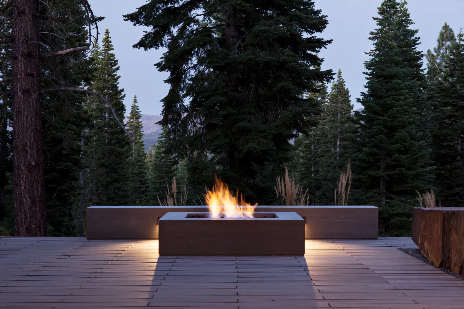 Modern mountain deck with a squared concrete lit fire pit at dusk. Surrounded by built-in bench seating and tall evergreen trees frame the space, with a distant forest.